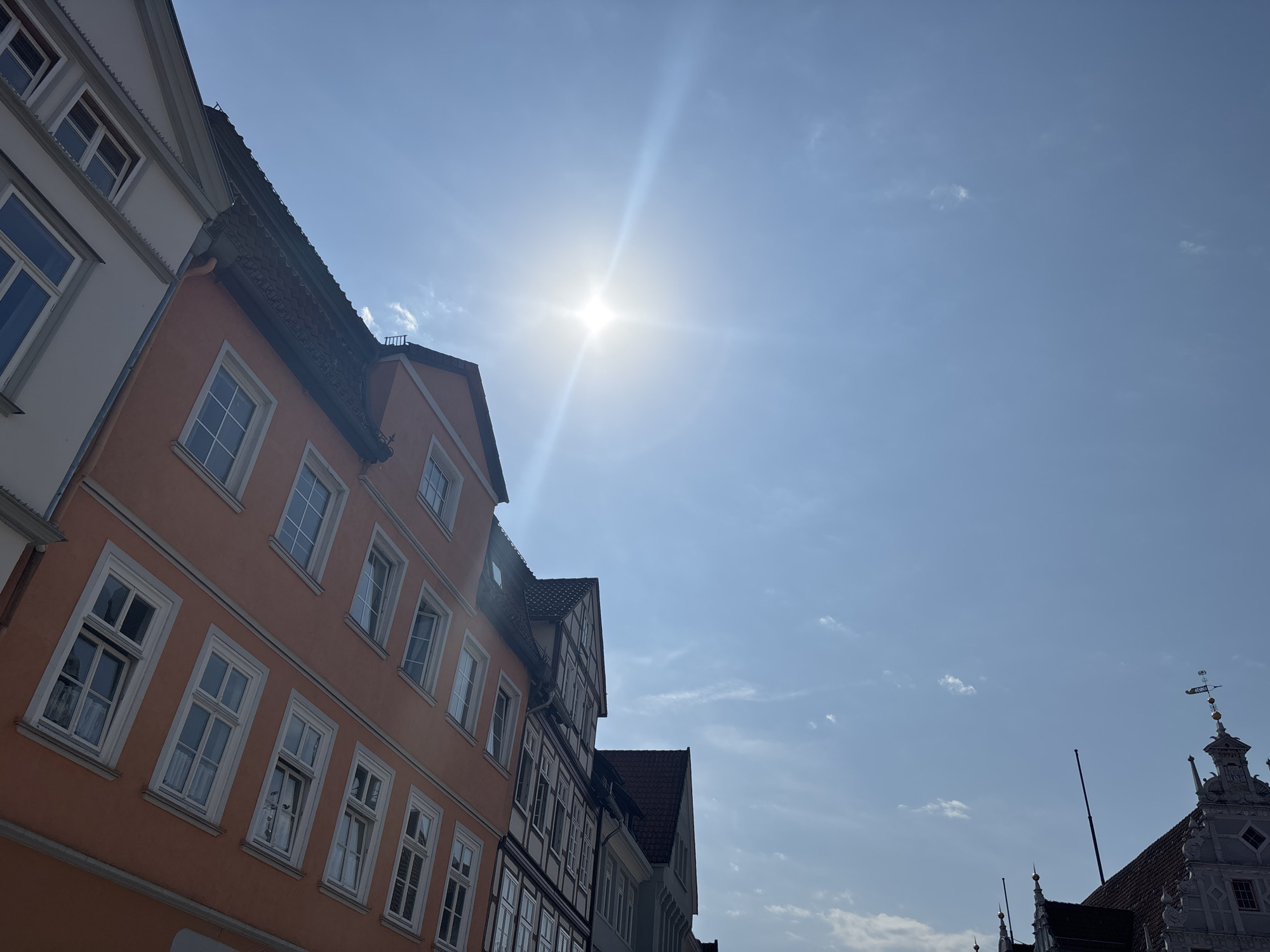 The image captures a street scene in Celle, Germany, focusing on the facades of historic buildings under a bright, sunny sky with scattered clouds. The sun is a prominent feature, emitting strong lens flare that radiates across the upper portion of the frame. The buildings are multi-story structures with traditional architecture. One building in the foreground is painted a vibrant orange and features numerous windows with white frames. To its left, another building is white with gray trim and also displays multiple windows. Further down the street, other buildings exhibit characteristic half-timbered construction with dark timbers and lighter infill. The rooflines are varied, with some featuring dark tiles and others showing lighter materials. In the far right of the image, a highly ornate building with decorative elements and a weather vane is visible, suggesting a significant historical structure. The perspective is angled upwards, emphasizing the architecture and the expansive sky. There are no people visible in the frame.