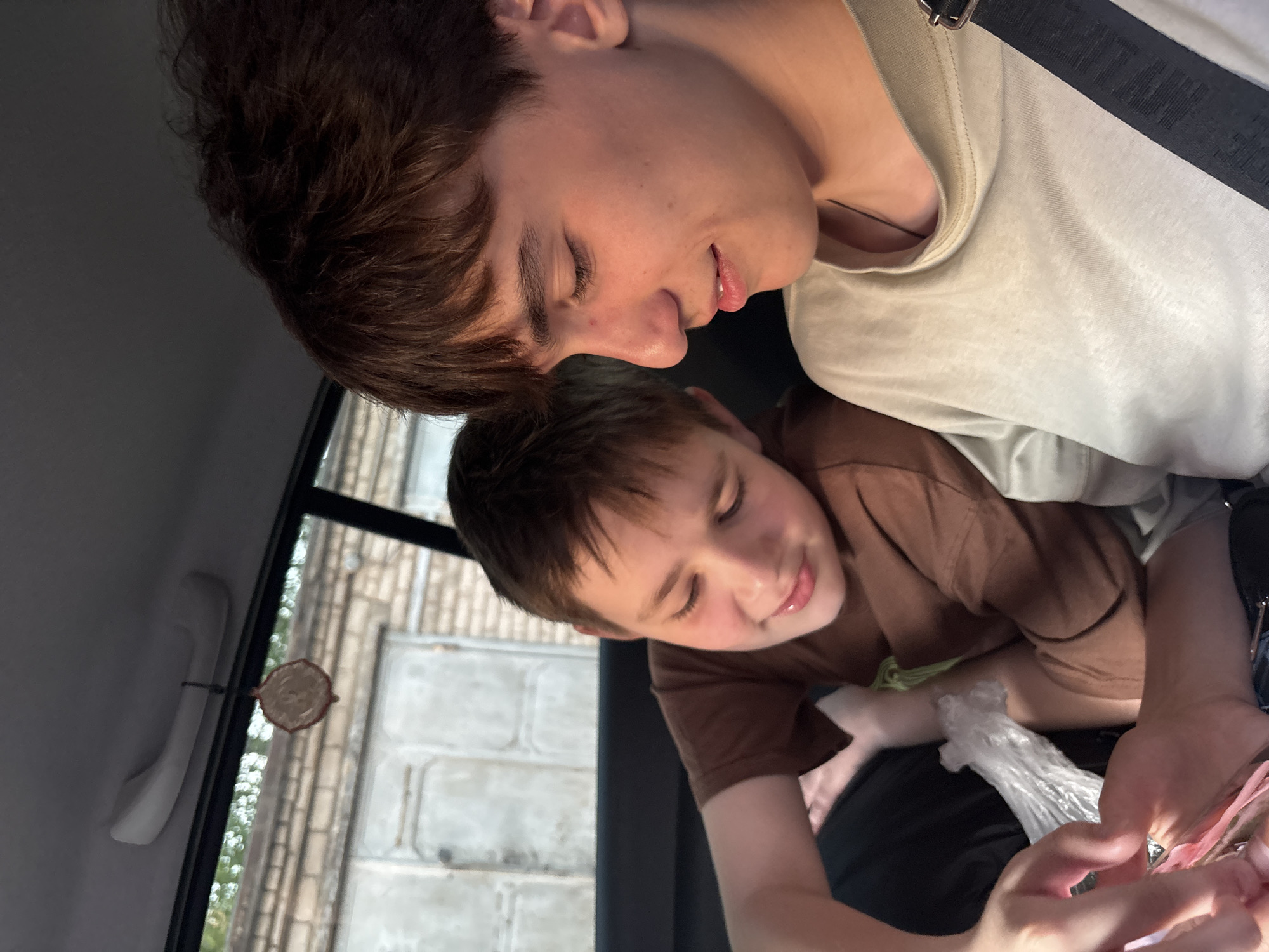 Two people are in a car. The older individual has dark brown hair and is wearing a light-colored t-shirt and a black shoulder strap. The younger individual has short brown hair and is wearing a brown t-shirt. A plastic bag is visible between their hands. The window shows a brick building exterior. A hanging car freshener is attached to the window handle. The location appears to be in an urban area with brick buildings, potentially matching building styles in Chaikovsky, Russia.