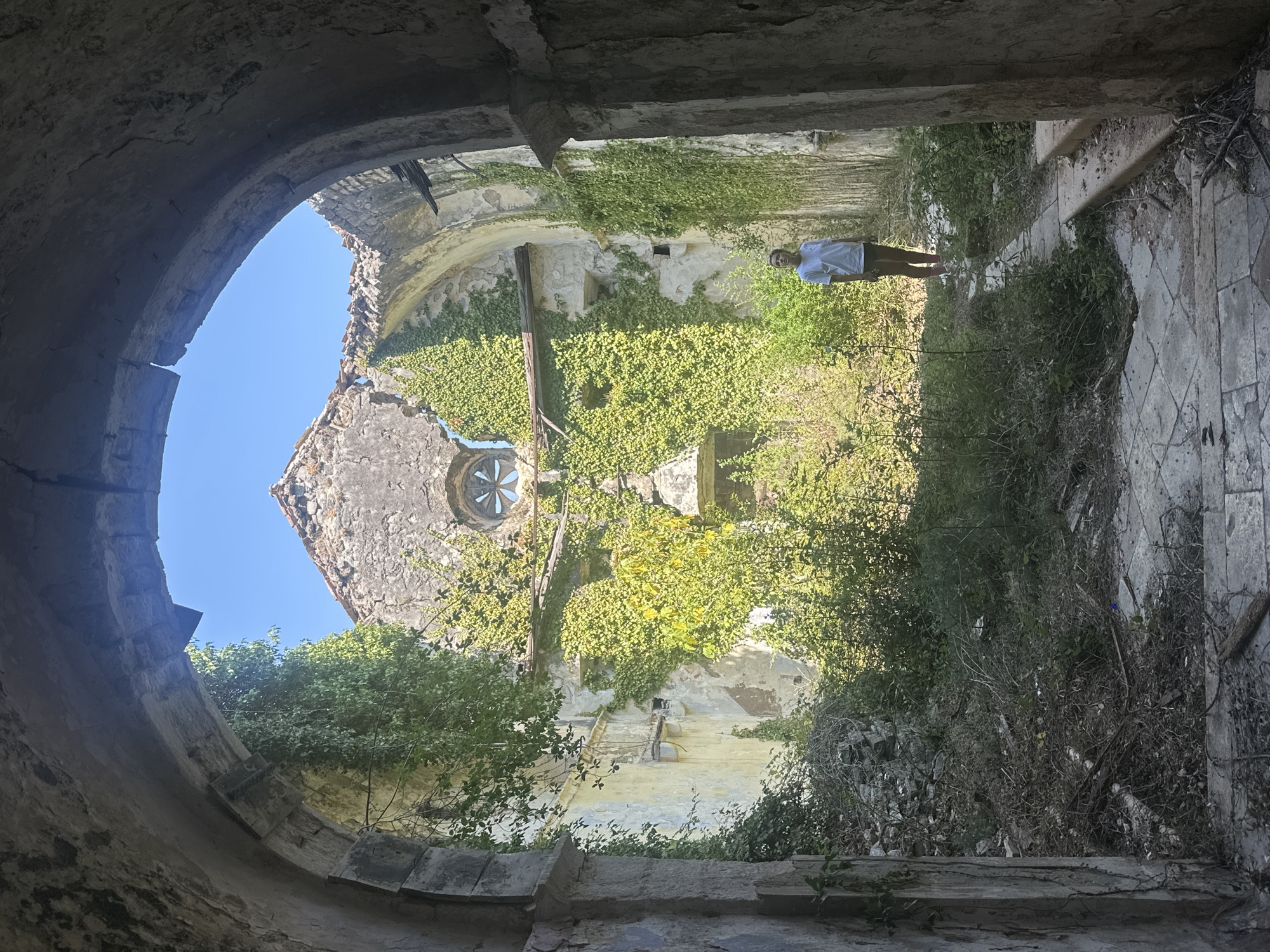 The image shows a person standing inside a ruined stone building in Prčanj, Montenegro. The building appears to be an old church or monastery, with crumbling walls and a partially collapsed roof open to the sky. Thick green ivy covers much of the interior walls and exterior facade. Sunlight streams in from the opening in the roof and through a circular window with a flower-like design. The floor is paved with large stone tiles, and debris, including branches and stones, is scattered around. The person is wearing a white t-shirt and dark shorts, and is looking towards the camera. The overall impression is one of decay and natural reclamation.