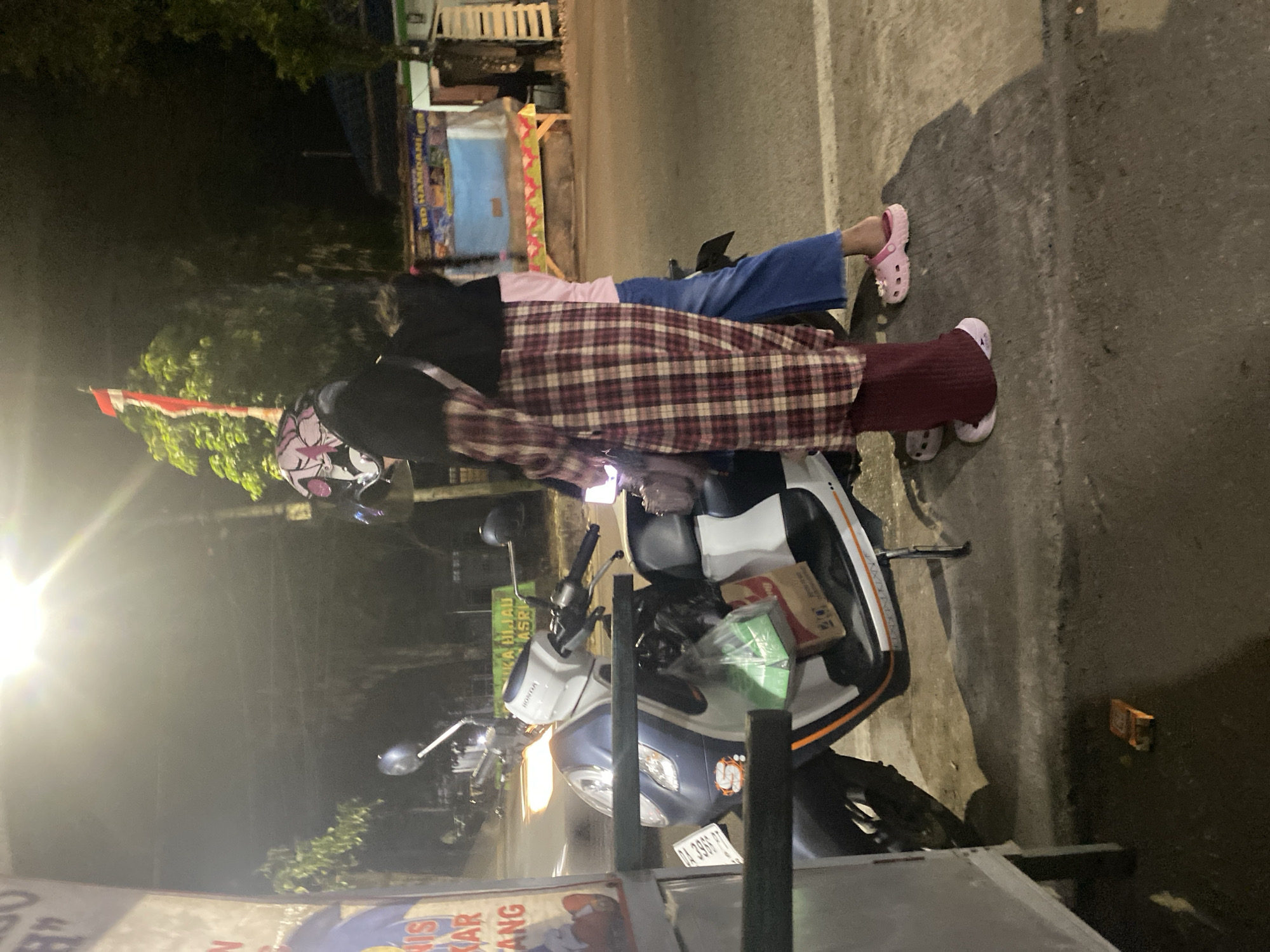 In Banjarbaru, Indonesia, at night, two individuals are standing next to a parked scooter. The scooter is white and blue, with a license plate that is partially obscured. The person on the left is wearing a black hijab and a patterned outer garment. The person on the right is wearing a pink shirt and blue pants. They are both wearing casual footwear. The scooter has items in its storage compartment, including a green bag and a cardboard box. In the background, there are trees, a building with a blue awning, and a street light. The scene appears to be on the side of a road, indicated by the asphalt surface and curb.