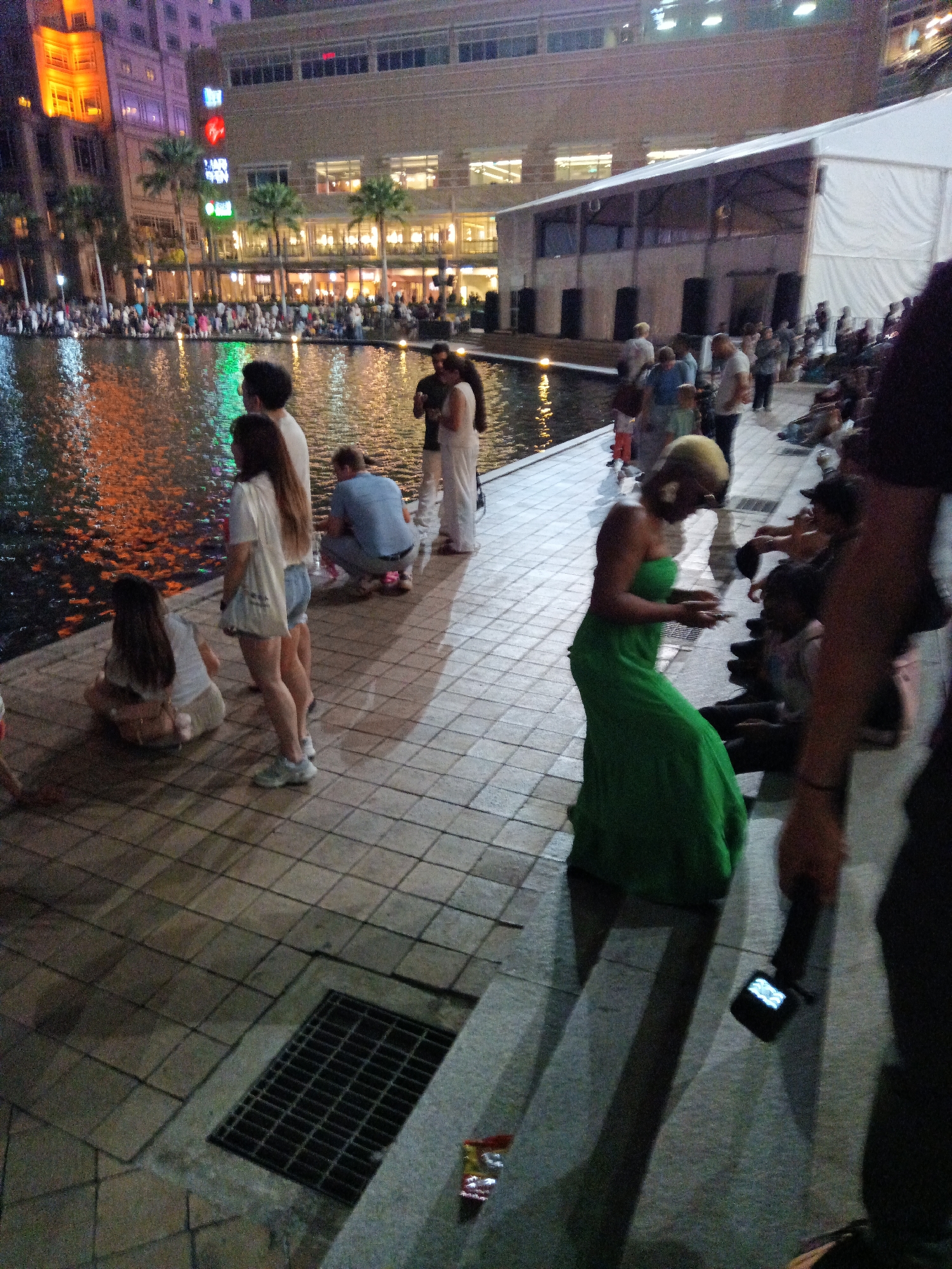 The image captures a lively outdoor scene at night, likely in Kuala Lumpur, Malaysia, given the presence of the distinctive "Virgin" logo which is prominent in the city. In the foreground, a woman in a green dress is standing and looking down at her phone. To her left, two individuals are seated on the paved ground, and behind them, a group of people are gathered by a body of water. The water reflects the colorful lights of the surrounding buildings, creating a vibrant and dynamic atmosphere. Further in the background, a large white tent structure is visible, possibly part of an event or exhibition. The architecture of the buildings suggests an urban setting, with modern structures and illuminated facades. The overall scene depicts a public gathering, with people enjoying the evening ambiance by the water.