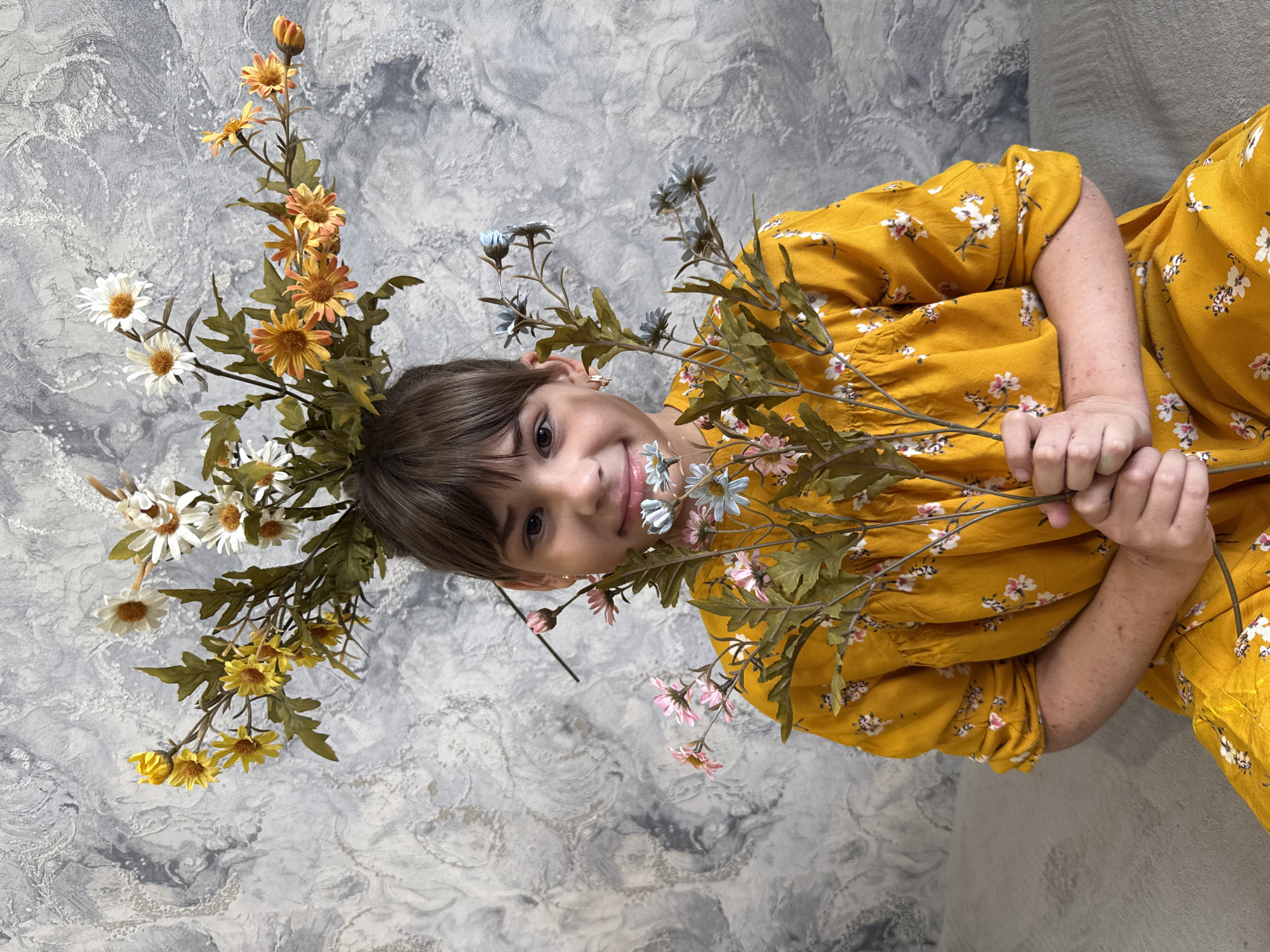 A child wearing a yellow, flower-patterned dress is lying on a gray surface, surrounded by artificial flowers. The flowers are arranged in a crown around the child's head and also held in the child's hands. The environment is undefined, seemingly indoors against a textured wall or backdrop. The location is Unknown, Unknown.