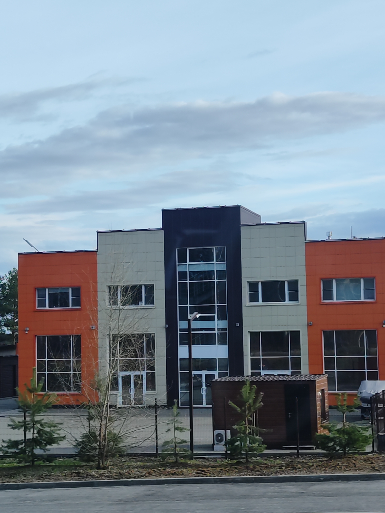 The image shows a modern commercial building located in Izhevsk, Russia. The building has three distinct sections: one painted orange, one beige, and a central section with black cladding featuring a glass facade. There are large windows on the ground and second floors. A small wooden shed is present in front of the building, along with young pine trees and a chain-link fence. The sky is overcast with clouds.