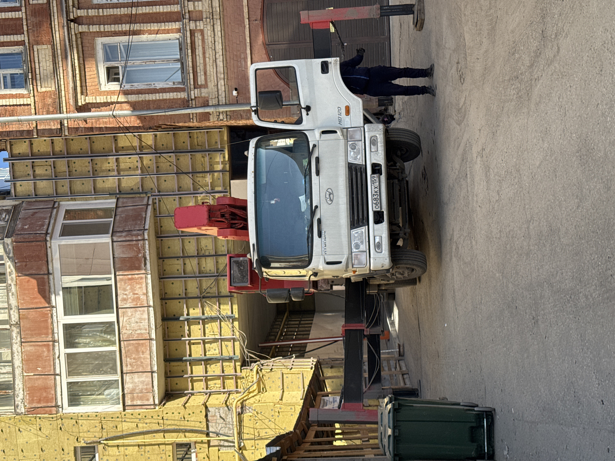The image captures an urban scene in Perm, Russia, during bright daylight, showing a construction or renovation activity. A white Hyundai HD120 truck, equipped with a red hydraulic crane or manipulator, is prominently positioned in what appears to be an alley or narrow street. The truck's license plate, "0683KX 159," is clearly visible, indicating its Russian origin. Its outriggers are extended, stabilizing the vehicle during operation.
A person, dressed in dark clothing, stands near the open driver's side door of the truck, seemingly involved in operating or attending to the crane.
To the left of the truck, a multi-story building is undergoing renovation. The upper part of the building features traditional red brickwork with white-framed windows. Below this, sections of the building are exposed, revealing yellow insulation material affixed to the facade, crisscrossed by metal framing in preparation for new cladding. Some areas show partially installed reddish-brown panels. The ground surface is paved concrete. The overall impression is one of active urban development or restoration work.
