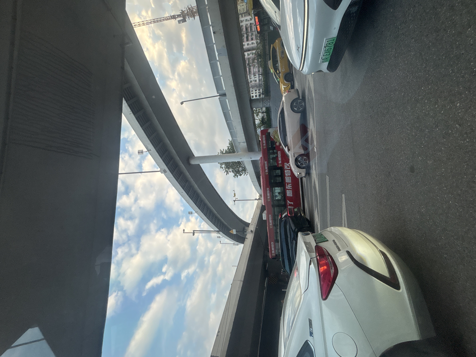 The image shows a street scene in Liwan District, China, with multiple overpasses creating a layered effect of infrastructure. A white car is in the foreground, partially obscuring a green license plate that reads "粤A EX6781". To the right, another white car is visible, with a portion of its front bumper and a similar license plate in view. In the middle ground, a red bus with Chinese lettering and a black SUV are present. Further back, a yellow taxi drives past. The sky is partly cloudy, with a bright sun casting light. A construction crane is visible in the distance on the right side of the image. The scene depicts a typical urban traffic environment with significant elevated roadways.