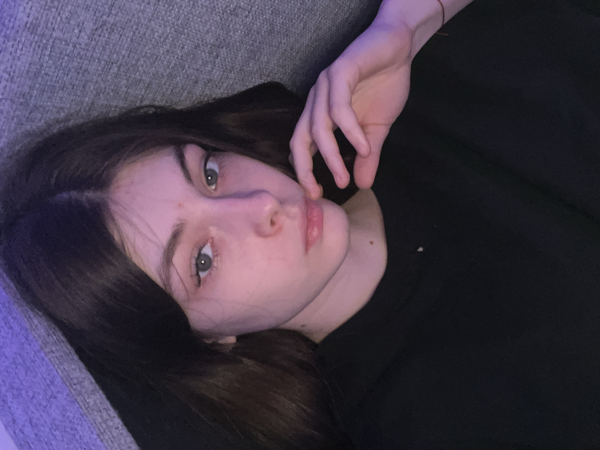 A young woman with long, dark hair lies on a gray fabric surface, possibly a couch or bed. She wears a black garment. Her head is tilted, and she looks directly at the camera with a somewhat contemplative expression. Her right hand is raised, with her fingers lightly touching her cheek. The environment appears to be indoors, possibly in a dimly lit room. The lack of details makes it impossible to determine the location in Saint Petersburg, Russia, or any other specific environmental details.