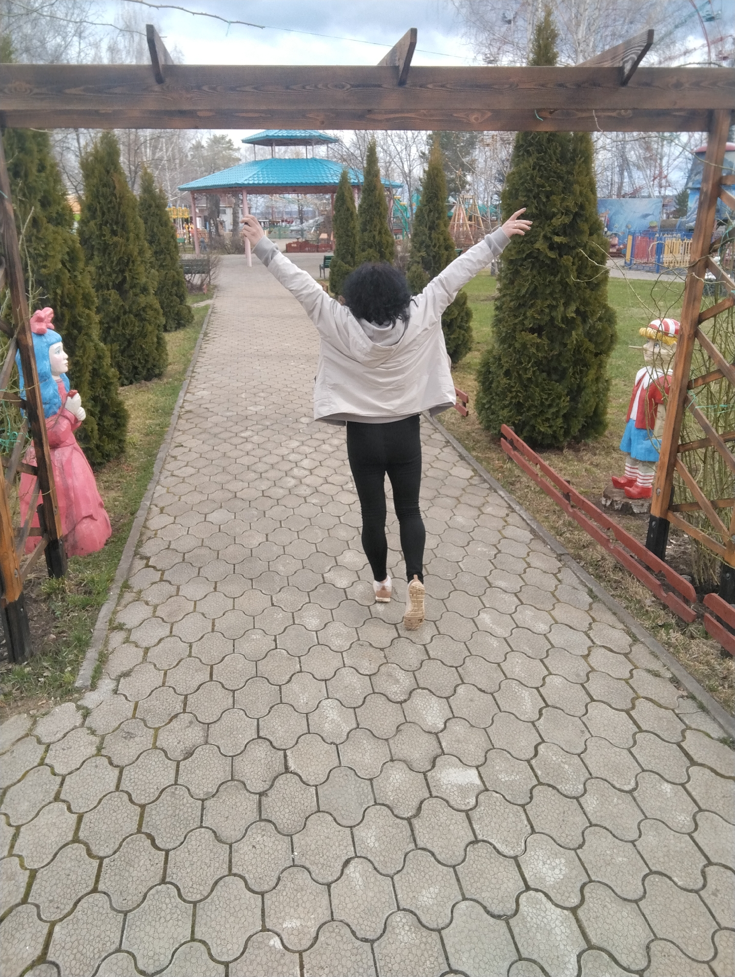 A full-length, rear view captures a person with dark curly hair walking down a paved pathway in what appears to be a park or amusement area in Chaikovsky, Russia. The person wears a light grey hooded jacket, dark leggings, and light-colored shoes, with their arms outstretched and raised, suggesting joy or freedom.
The pathway is made of light grey, interlocking concrete pavers with a distinctive wavy pattern, framed by green grass on either side. Tall, conical evergreen trees line both sides of the path, creating a verdant corridor.
At the immediate foreground, a rustic wooden archway or pergola frames the scene. Decorative figures are positioned near the archway on either side of the path. On the left, a statue resembling Alice from Wonderland, with blue hair, a pink bow, and a pink dress, holds a red object. On the right, a statue resembling Pinocchio, with a red and white striped hat, blue shorts, and red shoes, stands beside a short red wooden fence section.
In the background, beyond the trees, a blue-roofed gazebo or pavilion is visible, along with other amusement park structures, including what might be a Ferris wheel or similar ride, and colorful fences. Bare deciduous trees are also seen against a light, overcast sky, indicating possibly late winter or early spring. There is no visible text in the image. The overall atmosphere is one of leisure and enjoyment in an outdoor setting during daytime with mild weather.