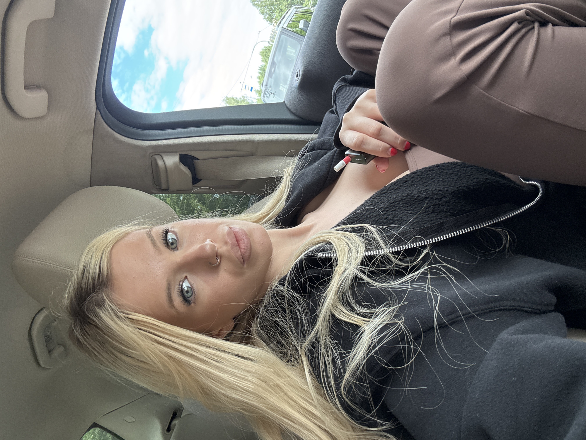 A blonde woman with blue eyes and a nose ring is sitting in the passenger seat of a car. She is wearing a black zip-up hoodie over a beige top and brown leggings. She is holding a vape device in her right hand. The car appears to be stopped, as evidenced by the trees and a traffic light visible outside the window. The sky is cloudy. The image does not provide any specific location information for Saint Petersburg, Russia.