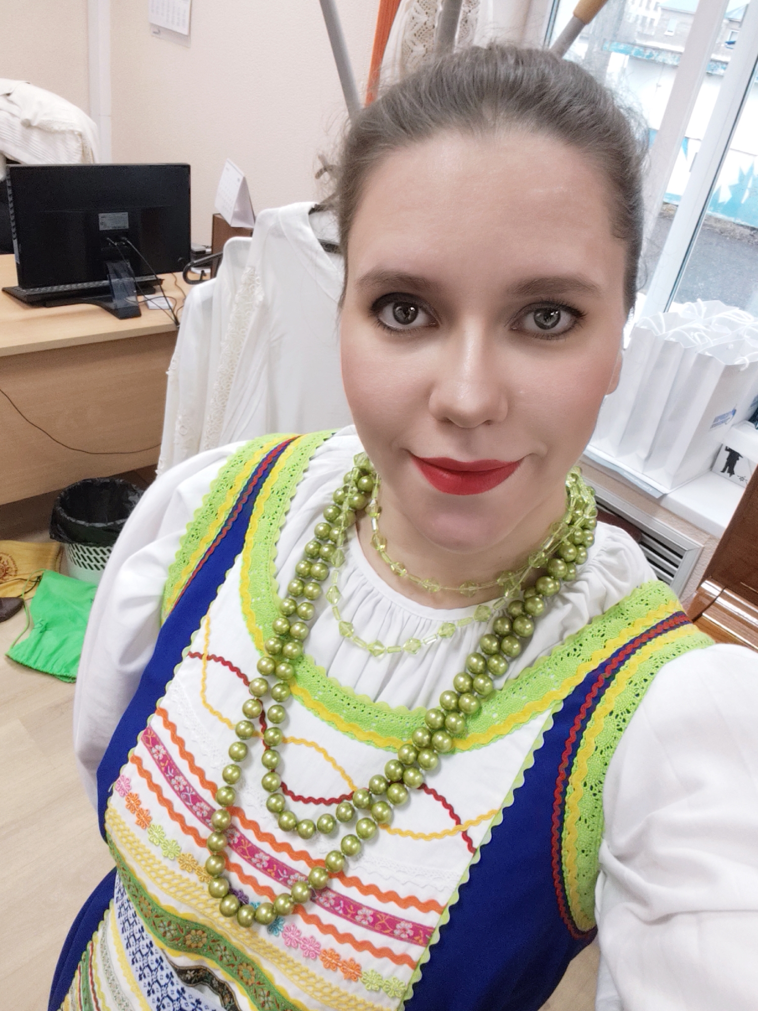 A woman is posing for a selfie in an indoor environment, seemingly in an office setting. She is wearing a traditional Russian dress with a white long-sleeved blouse, a blue vest with colorful embroidered patterns, and a lime green beaded necklace. Her hair is pulled back, and she is wearing red lipstick.
In the background, there is a desk with a computer monitor, a black trash can, and some white clothing hanging on racks. A window is visible, showing a blurred urban landscape. Based on the surroundings, it's likely she is in a work or studio space in an urban area. There is no definitive information about the location in Chaikovsky, Russia.
