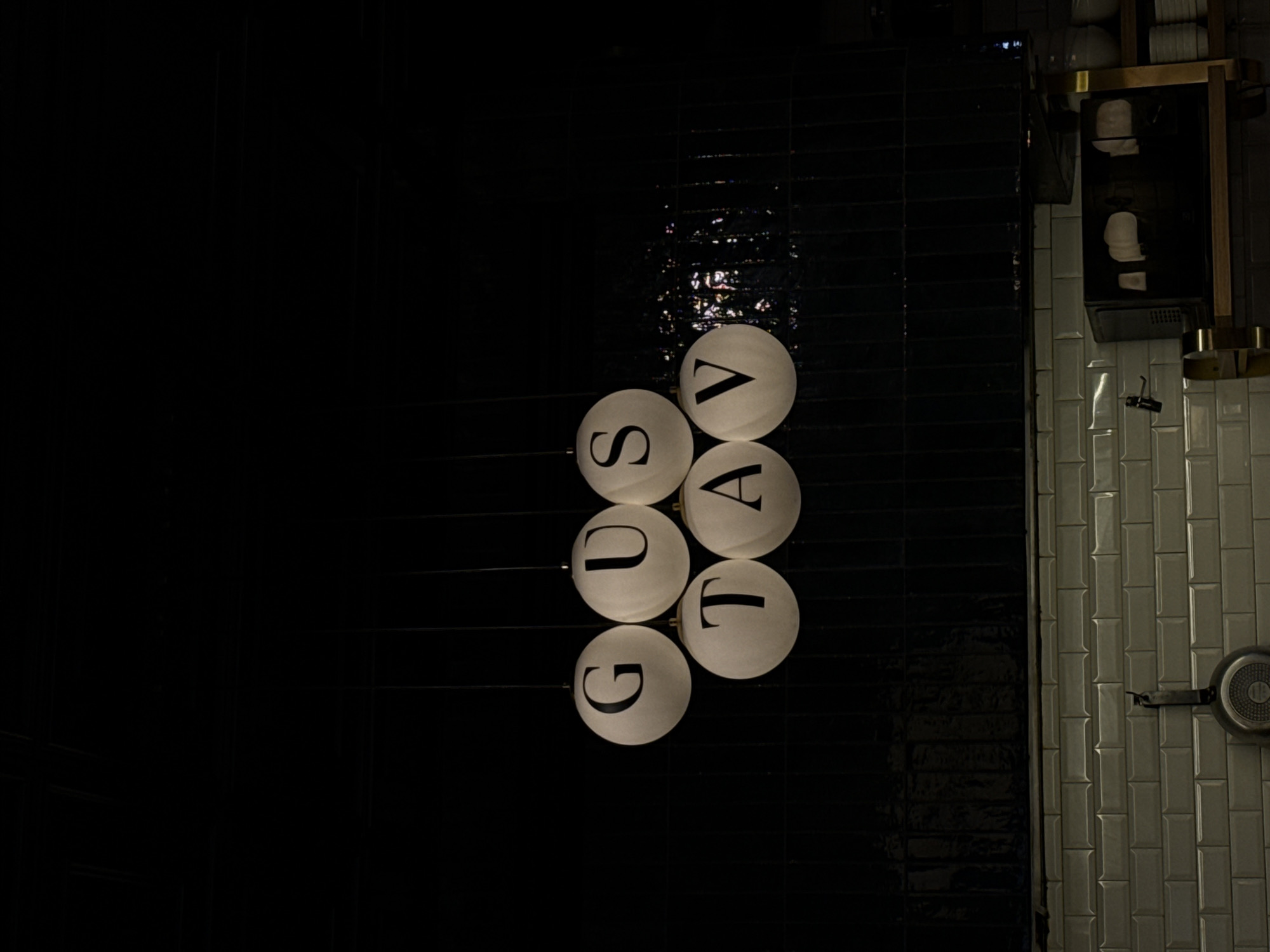 The image shows a close-up of a light fixture with the letters "GUSTAV" displayed on spherical lamps. The lamps are suspended from the ceiling against a dark, tiled background. Below the light fixture, a row of white subway tiles forms a backsplash, and a frying pan is hanging from a hook. To the right, a shelf displays stacked white bowls and a dark microwave. The setting appears to be a kitchen or dining area. The location is identified as Yerevan, Armenia.