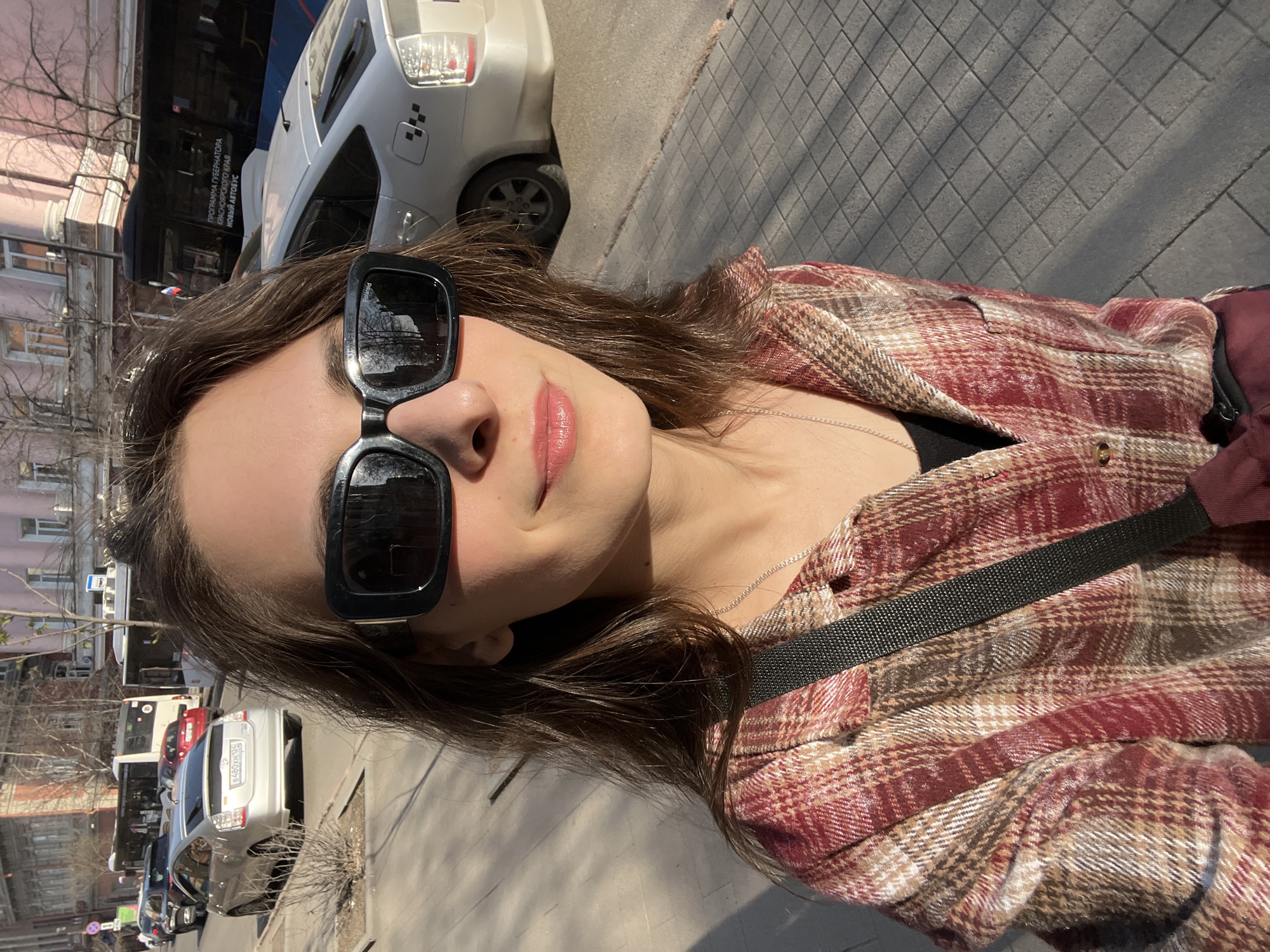 A woman is taking a selfie outdoors. She is wearing large black sunglasses and a red and brown plaid shirt. A thin silver necklace is visible around her neck. The background shows a street scene with cars and buildings. Based on the bus in the background that features a sign written in Russian that says “PROGRAMMA GUBERNATORA KRASNOYARSKOGO KRAYA - NOVYI AVTOBUS,” the location is Krasnoyarsk, Russia. The photo appears to be taken on a sunny day.