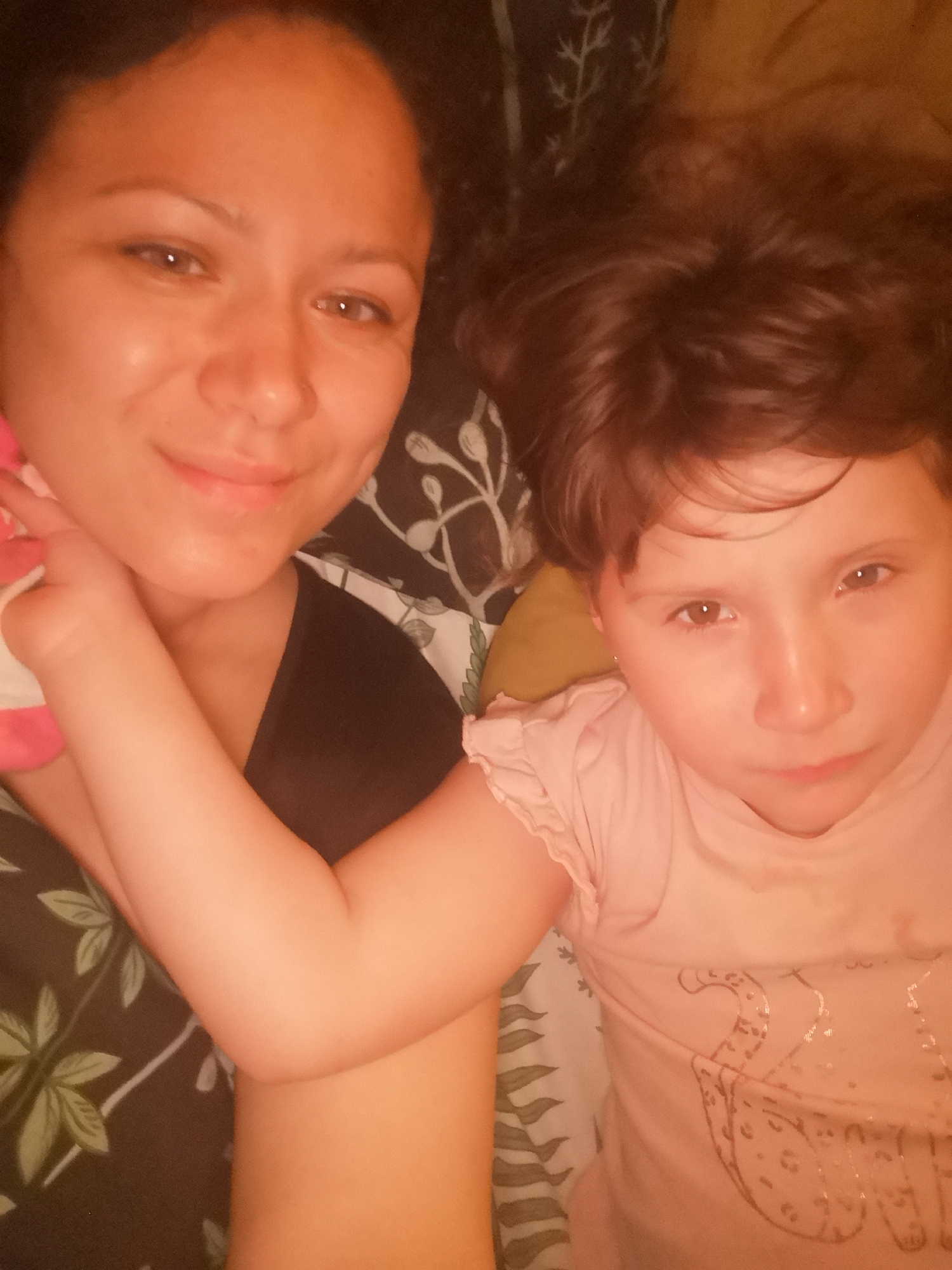 An overhead close-up shot shows a mother and daughter lying down together, with the mother on the left and the daughter on the right. The mother has dark hair and is wearing a black t-shirt. The daughter has brown hair and is wearing a pink t-shirt with a leopard print design. The daughter's arm is resting on the mother's chest, and the mother's arm is holding a pink toy. They are lying on a patterned blanket. The lighting is warm and suggests an indoor setting, possibly a bed.