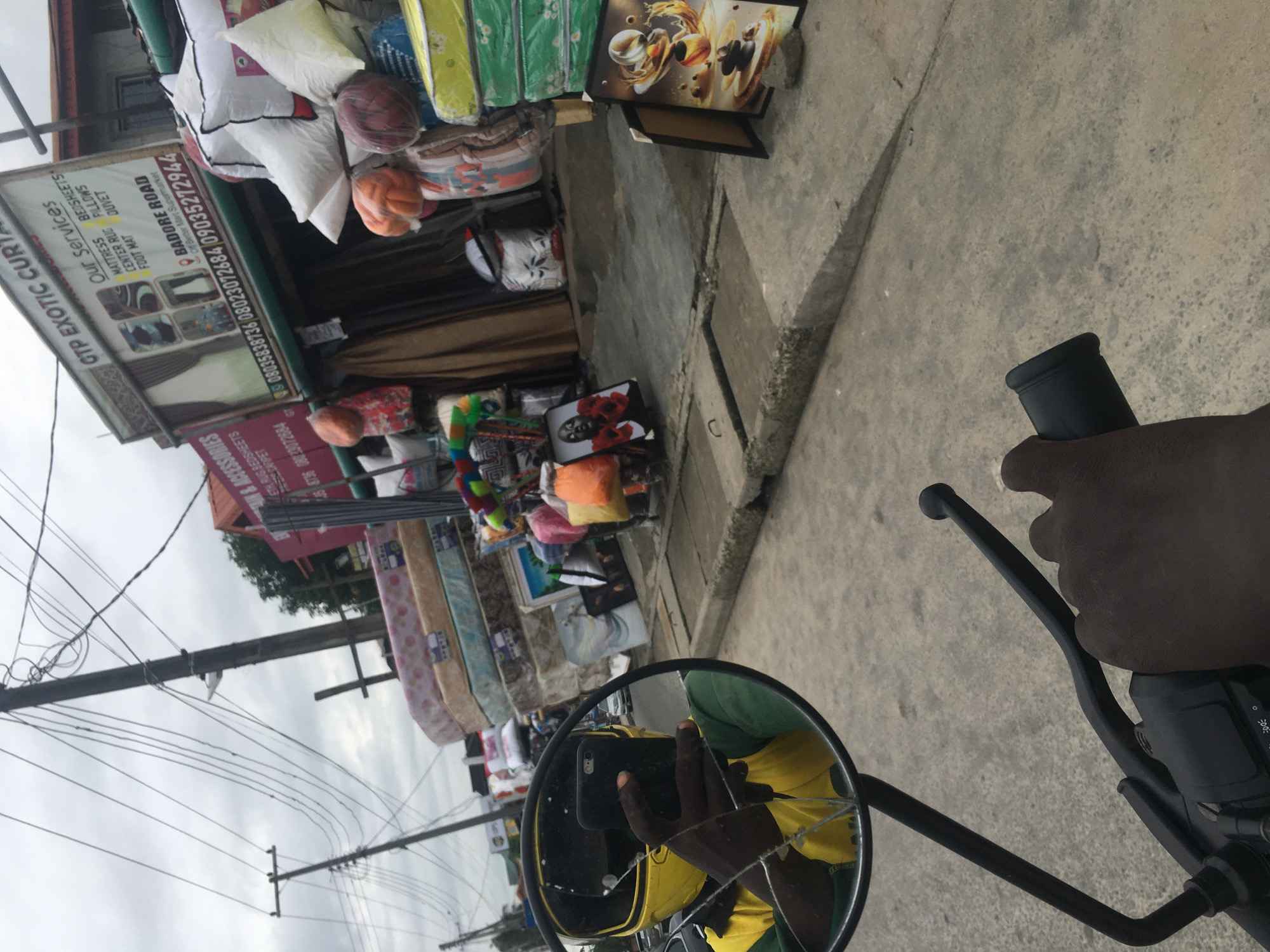 The image, seemingly taken from the perspective of someone on a motorcycle, shows a street-side shop specializing in bedding and textiles. The shop displays a variety of mattresses, pillows, rugs, and bedsheets. The shop's sign indicates that it is located on Badore Road, near the Better Mart Supermarket, in Ado, Nigeria. The camera is reflected in the broken rearview mirror. The hand of the motorcycle rider is visible. Power lines cross the sky.