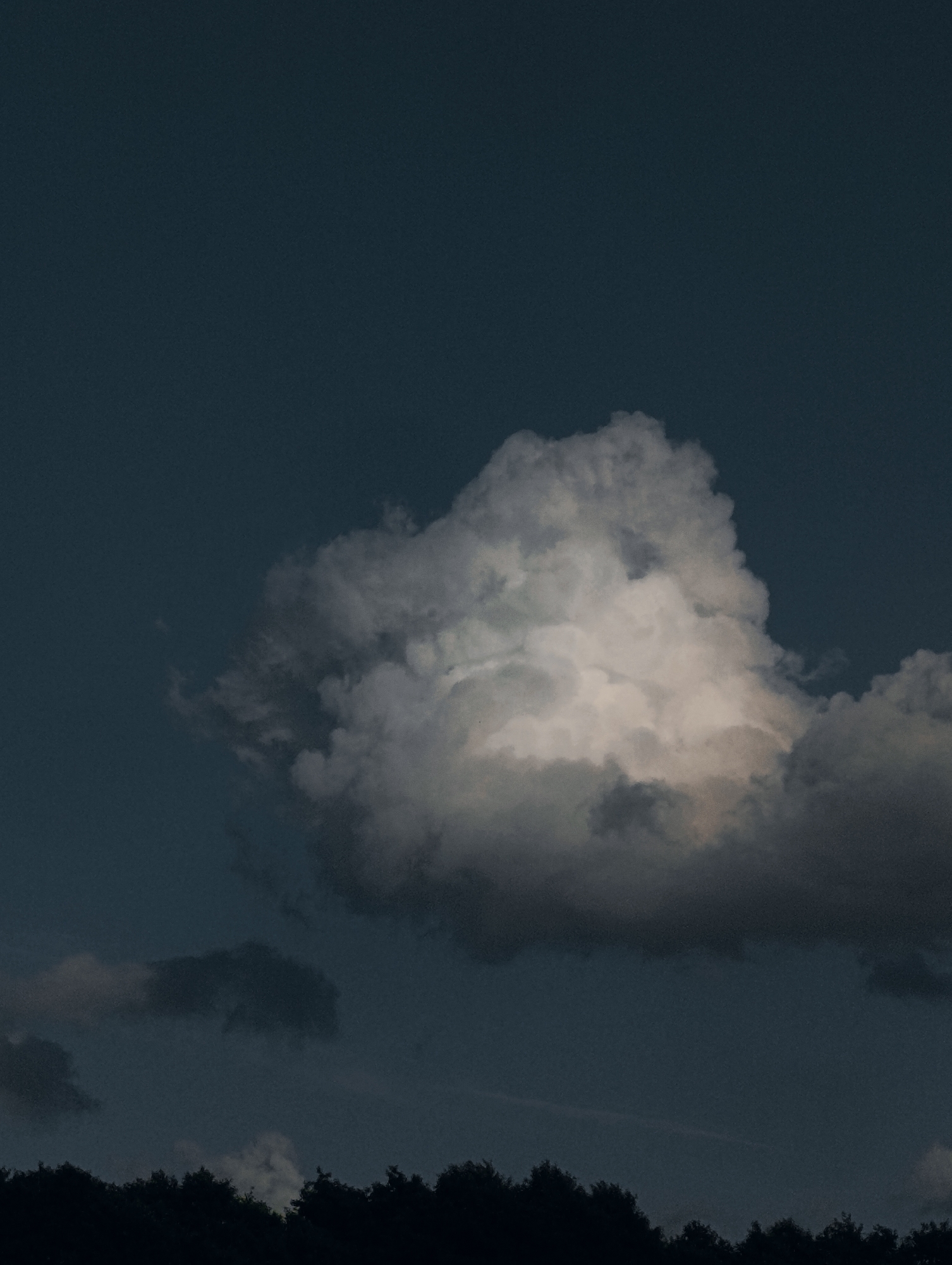 An image captures a single, luminous cloud against a deep twilight sky. The cloud is illuminated from within, casting a soft glow that contrasts with the darker, diffused clouds and the silhouetted tree line below. The overall scene is tranquil and atmospheric, with the dark blues and grays evoking a sense of calm evening or impending night. The location is unknown.