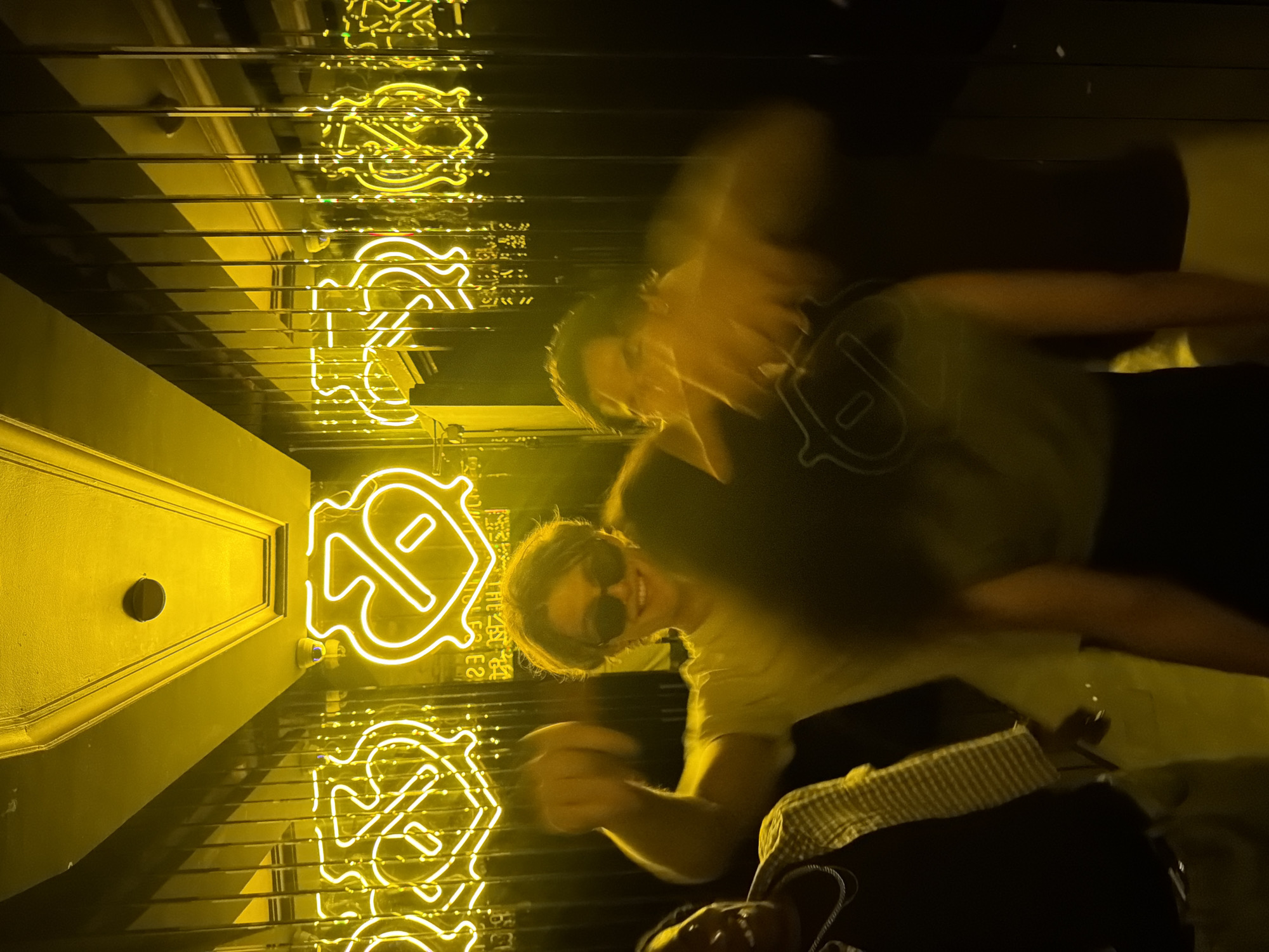 In Marrakesh, Morocco, four individuals are captured in a dimly lit environment, bathed in the vibrant yellow glow of neon signage. The signage, featuring a heart-shaped logo with a diagonal bar, is prominently displayed on a mirrored wall, creating a reflective effect. Two individuals are in the foreground. On the left, a person wearing a black t-shirt and a yellow and white checkered jacket, adorned with a silver chain, is visible from the chest up. In the center, a person with sunglasses and a light-colored t-shirt smiles broadly, with their right arm raised and fist clenched, as if in a gesture of excitement or celebration. Behind them, two other individuals are present but less distinct due to motion blur. The overall atmosphere appears to be one of revelry or a social gathering, likely within a club or a similar entertainment venue.