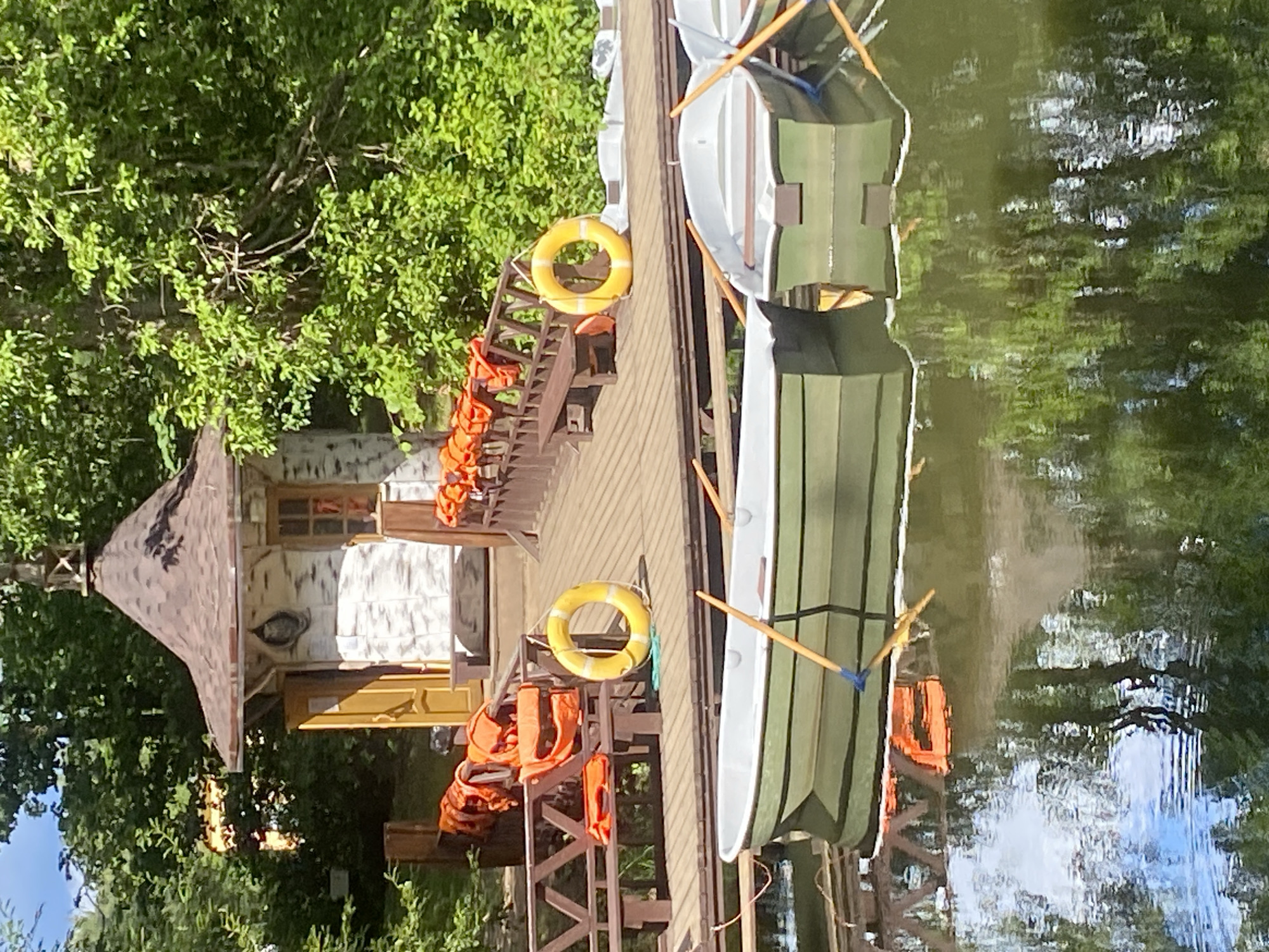 The image shows a scenic view of a body of water with several rowboats docked at a wooden pier. A small, charming building with a distinctive hexagonal roof stands on the pier, adorned with what appears to be birch-like siding. Orange life vests are neatly stacked on a railing along the pier, and a yellow life preserver ring hangs on a post. Lush green trees and foliage surround the area, creating a tranquil, natural setting. The reflection of the trees and sky on the water's surface adds to the picturesque ambiance. This scene is likely located within the city of Pavlovsk, Russia, a place known for its beautiful parks and palaces.