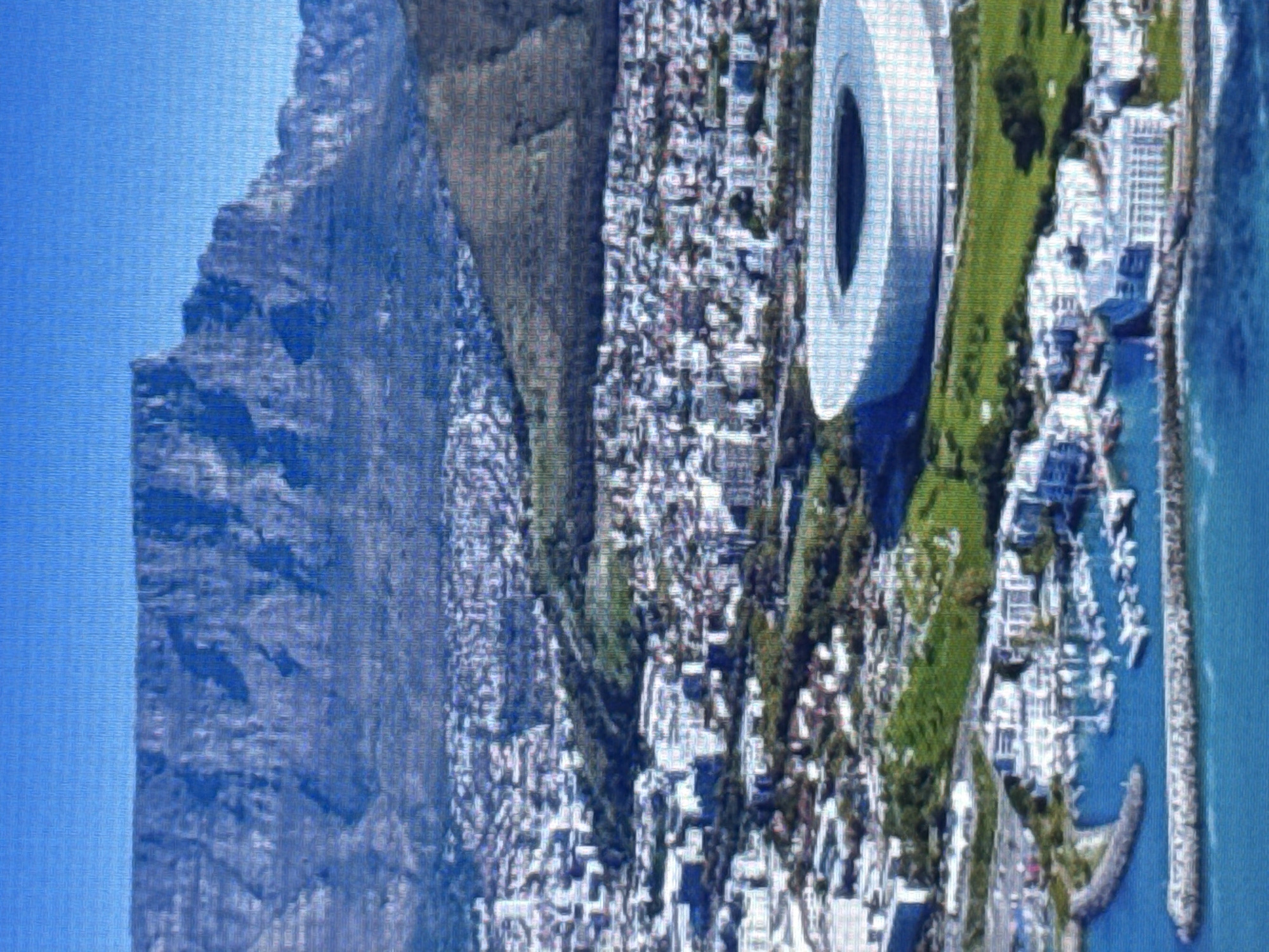 The image displays an aerial view of a coastal city nestled against a large, rugged mountain. The mountain dominates the left side of the frame, its steep slopes covered in rocky terrain. To the right of the mountain, a dense urban area stretches out, characterized by numerous buildings of varying sizes and architectural styles. Green spaces, likely parks or manicured lawns, are interspersed within the city.
A prominent circular stadium, with a white facade and a dark, open center, is a striking feature in the cityscape. Adjacent to the urban area, a harbor or marina is visible, filled with many white boats docked alongside piers. The water in the harbor is a calm, blue hue. Further out, the ocean extends to the right of the image, appearing clear and blue.
The scene suggests a sunny day, with bright lighting casting shadows and highlighting the details of the buildings and landscape. The time of day appears to be daytime, likely afternoon, given the quality of light. The combination of the mountain, the city, and the harbor strongly suggests a coastal location with significant elevation changes. There is no visible text in the image. The overall impression is one of a vibrant city situated in a dramatic natural setting.