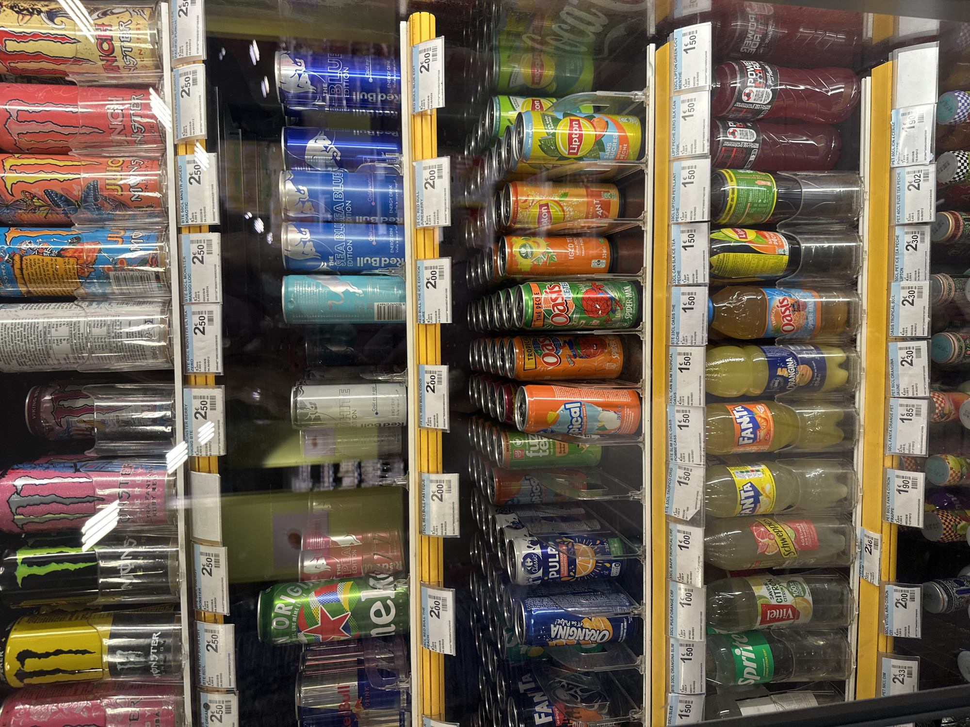 The image shows a refrigerated display case in a retail setting, likely a convenience store or supermarket, in Strasbourg, France. The shelves are stocked with a variety of canned and bottled beverages. Prominently displayed are energy drinks such as Monster Energy in various flavors, Red Bull in several editions, and other brands. Below these, there are selections of iced teas, fruit juices, and sodas, including Fanta, Sprite, Orangina, and Oasis. The lighting is bright, reflecting off the glass doors of the display. Price tags are visible for many of the products. The overall scene is clean and orderly, typical of a retail environment. There are no people visible in the image. The contents of the display indicate a focus on chilled drinks.