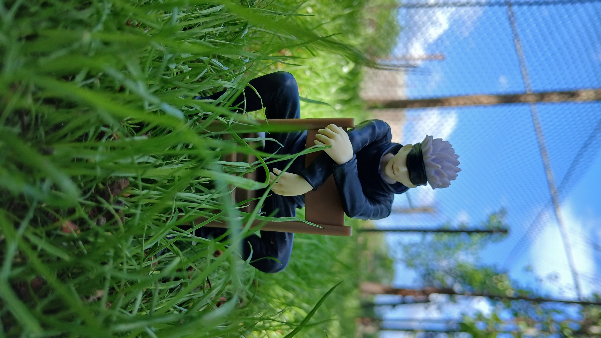 A figure of a seated anime character, likely Satoru Gojo from Jujutsu Kaisen, is positioned within a patch of green grass. The figure is seated in a brown miniature chair, wearing a dark outfit and a blindfold. The background shows a blurred view of a chain-link fence, trees, and a blue sky with white clouds. The location is not specified.