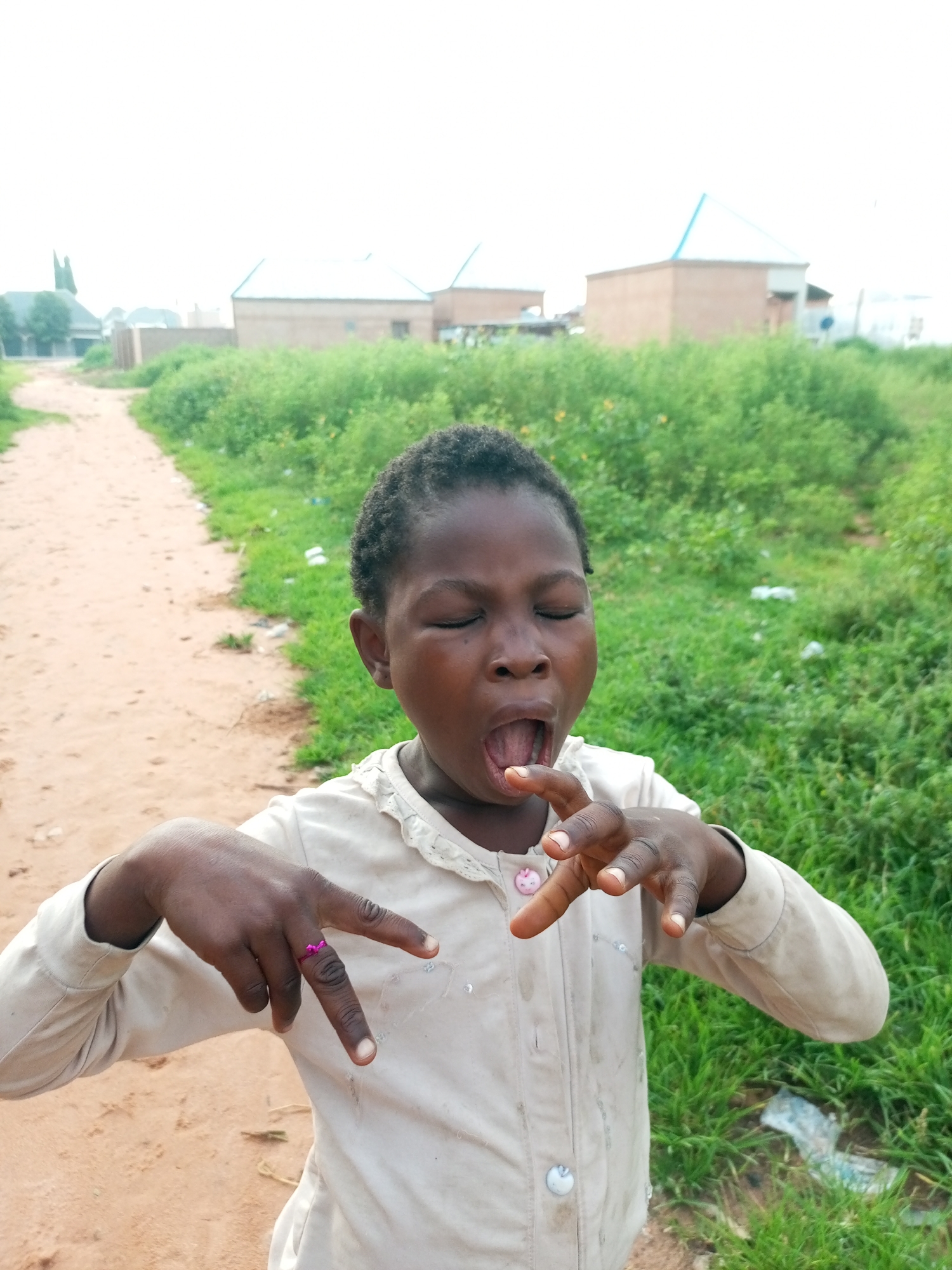 The image shows a young Black child with their eyes closed and mouth open, appearing to be singing or shouting. The child is wearing a light-colored, long-sleeved shirt with decorative buttons. The child's left hand, wearing a pink ring, is raised towards the camera, with fingers extended. The child is standing outdoors on a dirt path, with lush green grass and vegetation to the right. In the background, there are several buildings with brick walls and light-colored roofs, suggesting a residential area. The sky is overcast. This scene is located in Tumfure, Nigeria.