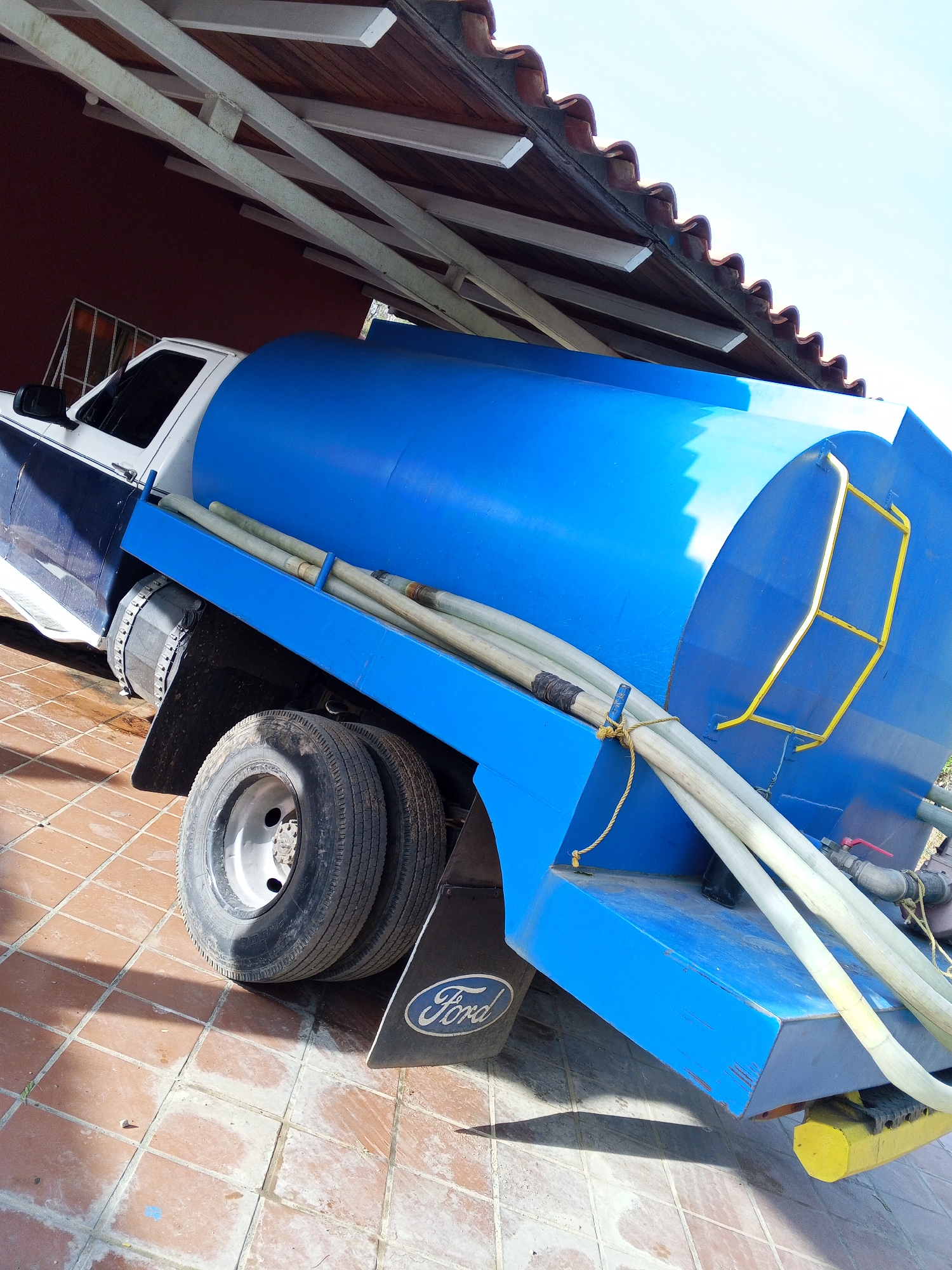 A blue water truck with a Ford mud flap is parked on a tiled surface under an overhang in Unknown, Unknown. The truck has a white cab with a blue stripe and a large blue tank. Several hoses are connected to the back of the truck. The overhang is supported by white beams and has a red tile roof. The sky is visible in the background.