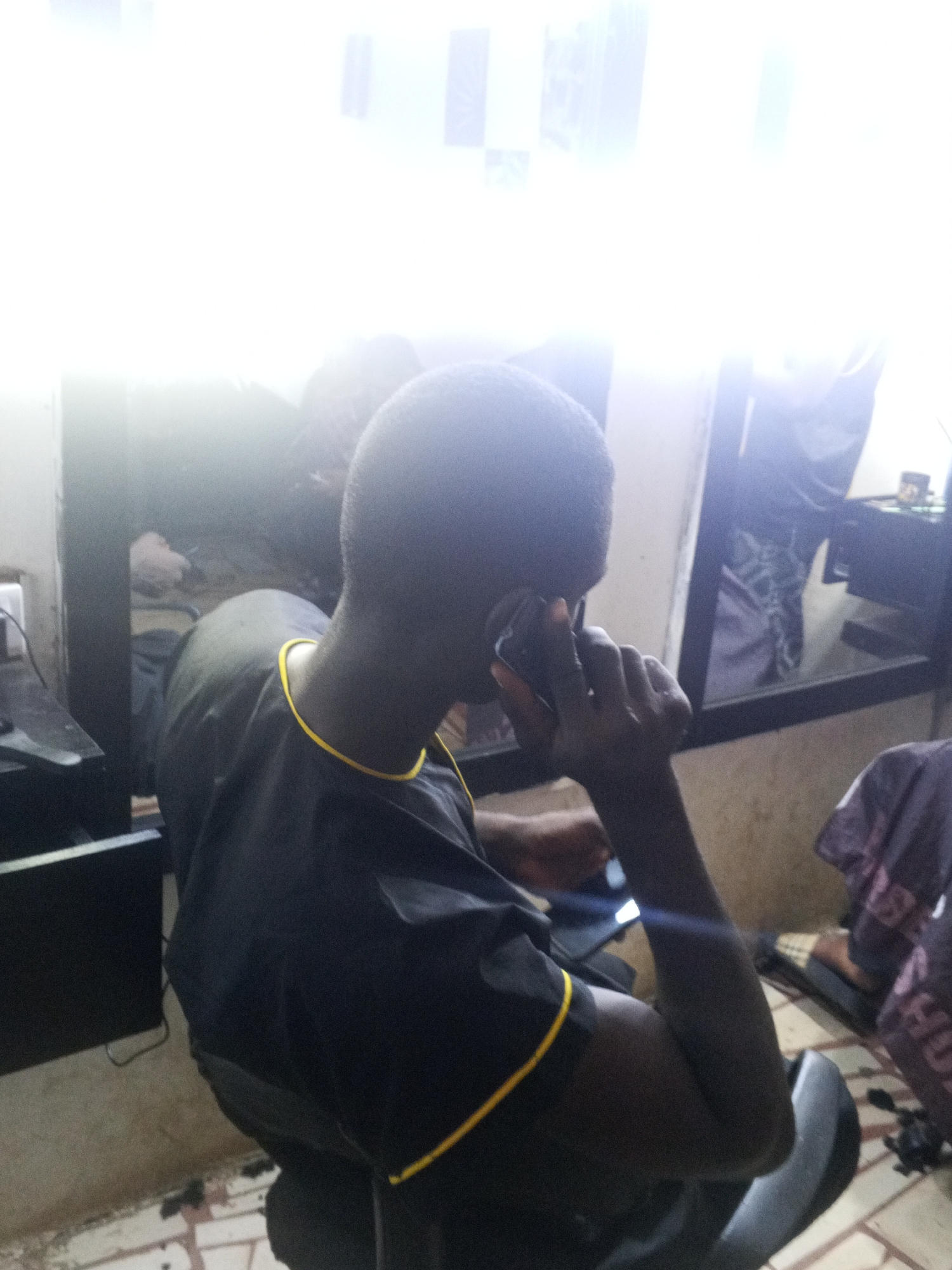 A person is sitting in a chair in what appears to be a barber shop in Gwong, Nigeria, talking on a cell phone. They are wearing a black gown with yellow trim around the neck and arm openings. A mirror is visible behind them, reflecting the surroundings. Hair clippings are scattered on the tiled floor.