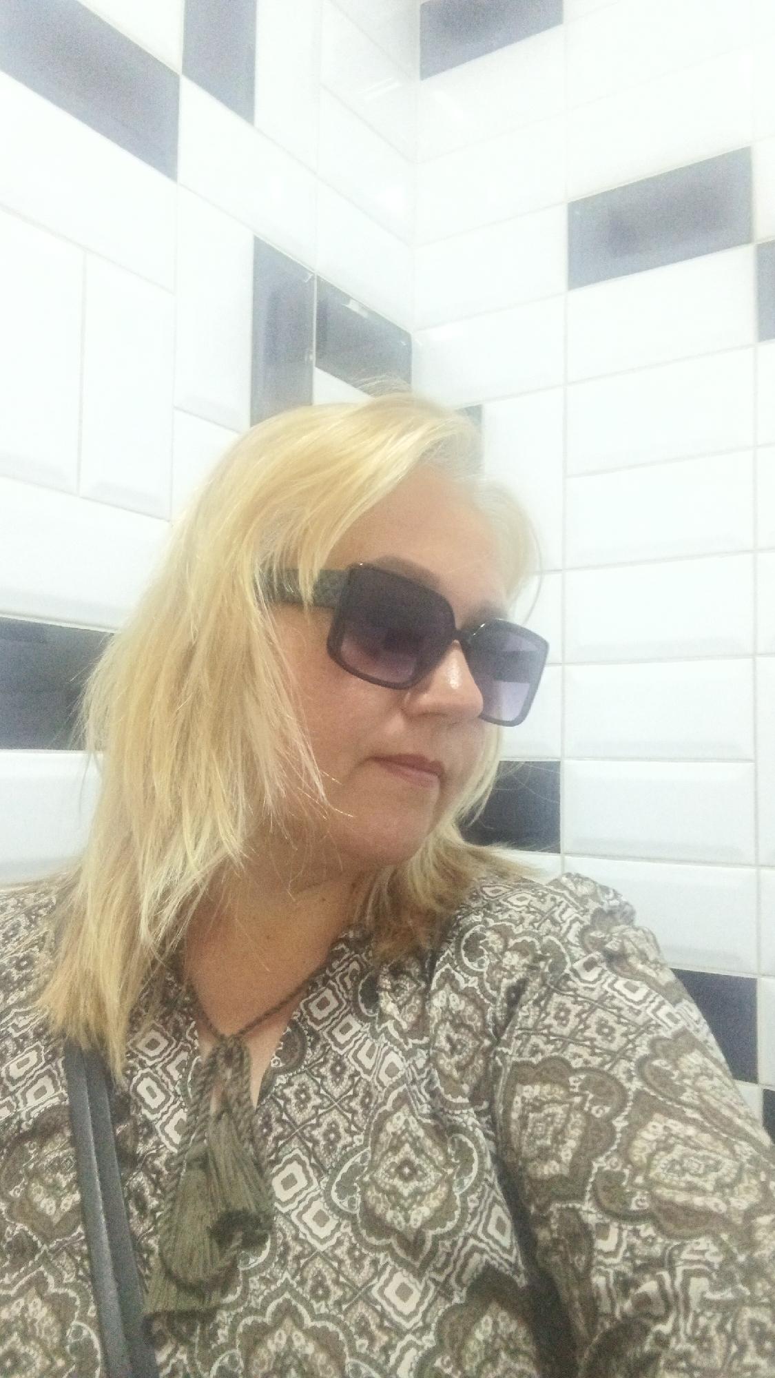 A woman with blonde hair and large sunglasses is shown from the chest up. She is wearing a patterned shirt with a tassel necklace. The background consists of white and black tiles, suggesting an indoor setting such as a bathroom or shower stall. The image is a selfie taken from a low angle. The location in Kryvyi Rih, Ukraine is not discernible from the image.