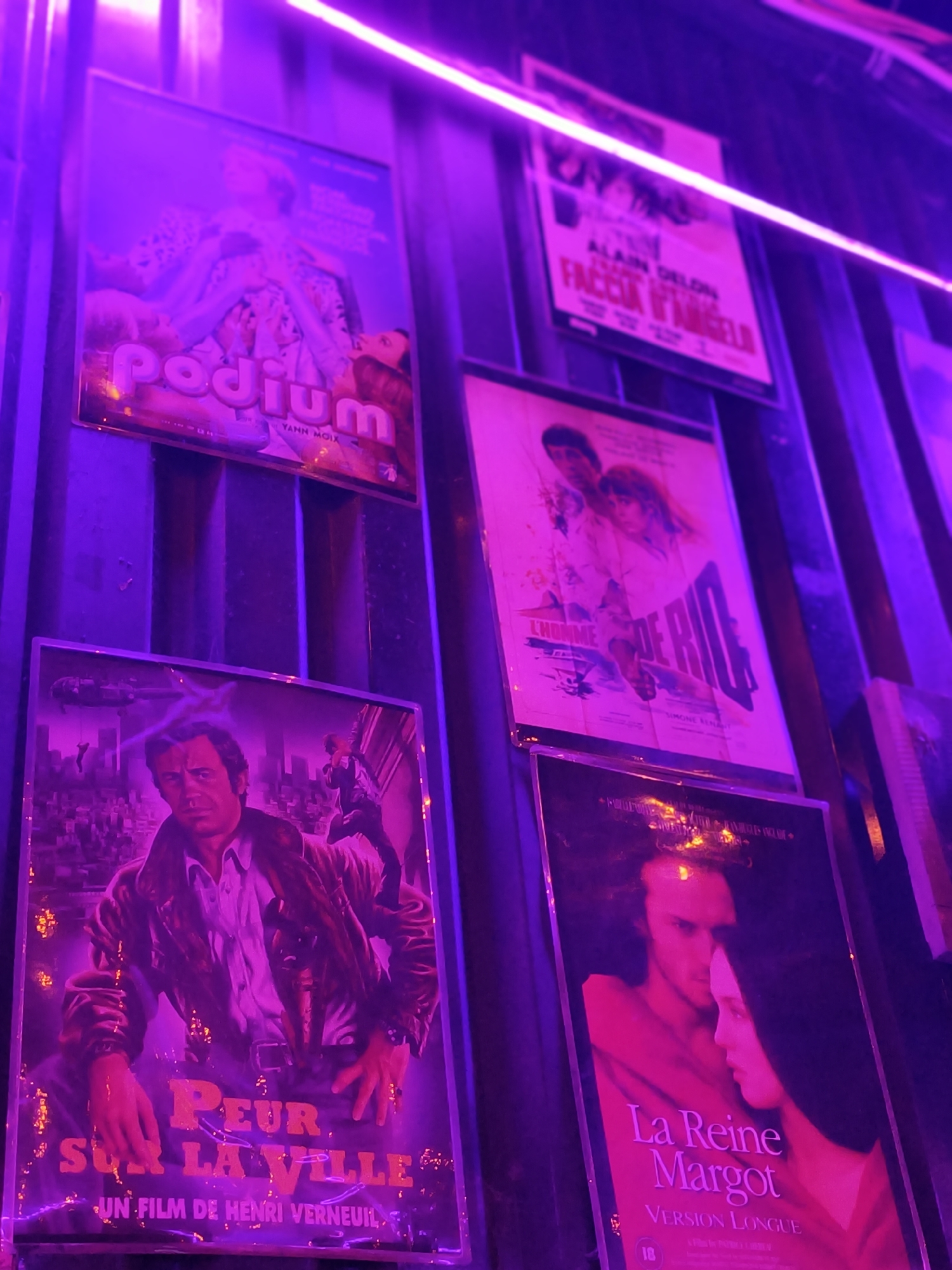 The image shows a collection of movie posters displayed vertically on a wall, illuminated by purple neon lights. The posters are in varying degrees of focus, with some sharper than others.
The top left poster features the title "Podium" in large, stylized font. Below it, smaller text reads "Yann Moix." The image on this poster appears to depict a person in an elaborate outfit.
To the right of "Podium" is another poster with French text, including "ALAIN DELON" and "FACCIA D'ANGELO." The image on this poster is mostly obscured by the neon lights and blur.
Below the "Podium" poster is a poster for "PEUR SUR LA VILLE," featuring a close-up of a man's face and upper body. The title is prominently displayed in large letters, and "UN FILM DE HENRI VERNEUIL" is visible at the bottom.
To the right of "Peur sur la Ville" is a poster for "LA REINE MARGOT," showing a man and a woman's profiles facing each other. The title "La Reine Margot" is in pink lettering, and "VERSION LONGUE" is below it.
The wall behind the posters appears to be made of vertical panels. The overall lighting suggests a nighttime or dimly lit indoor environment, possibly a cinema lobby or a themed bar. There are no people clearly visible interacting with the posters. The location is not identifiable as being in the city of Đà Nẵng, Vietnam, based on the visual information provided.