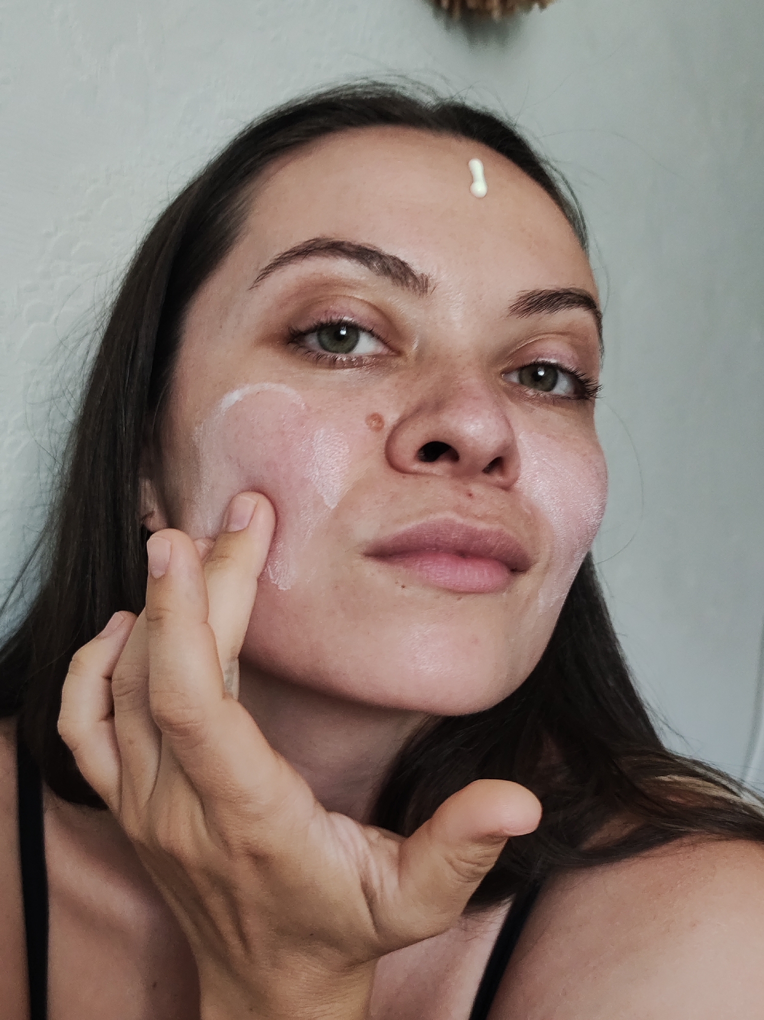 A woman with olive skin and dark hair is applying white cream to her face. There is a blob of cream on her forehead. She is looking at the camera and smiling slightly. Her left hand is visible in the foreground. She appears to be in an indoor environment with a light-colored wall in the background. There is no information visible in the image that would place this scene in the city of Chaikovsky, Russia, or any other specific location.