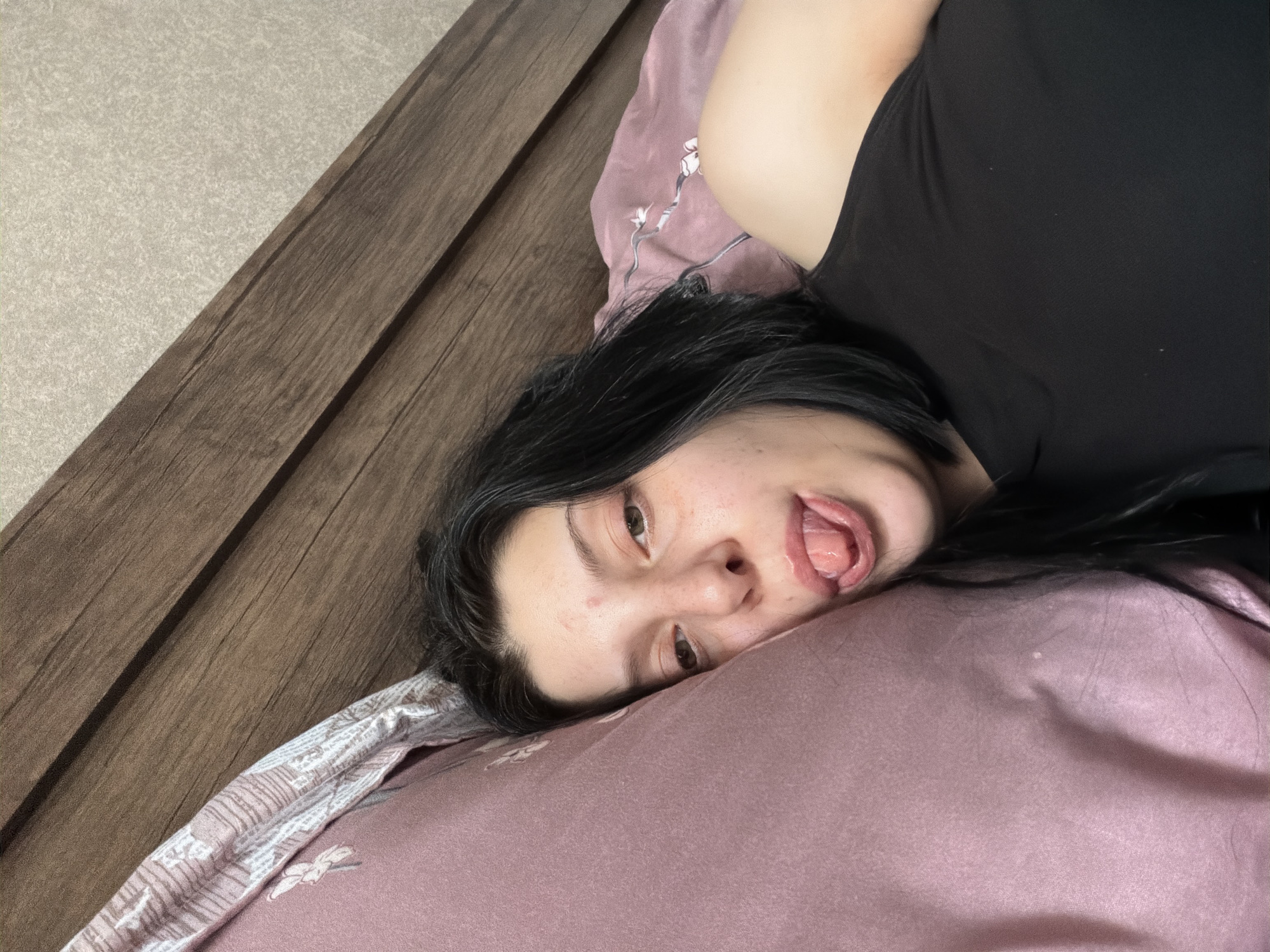 A young woman with dark hair and fair skin is lying on a bed, propped up against a pink pillow. Her eyes are open, and she is sticking her tongue out. She is wearing a black top. The background consists of a wooden headboard and a textured wall. The setting is a bedroom.