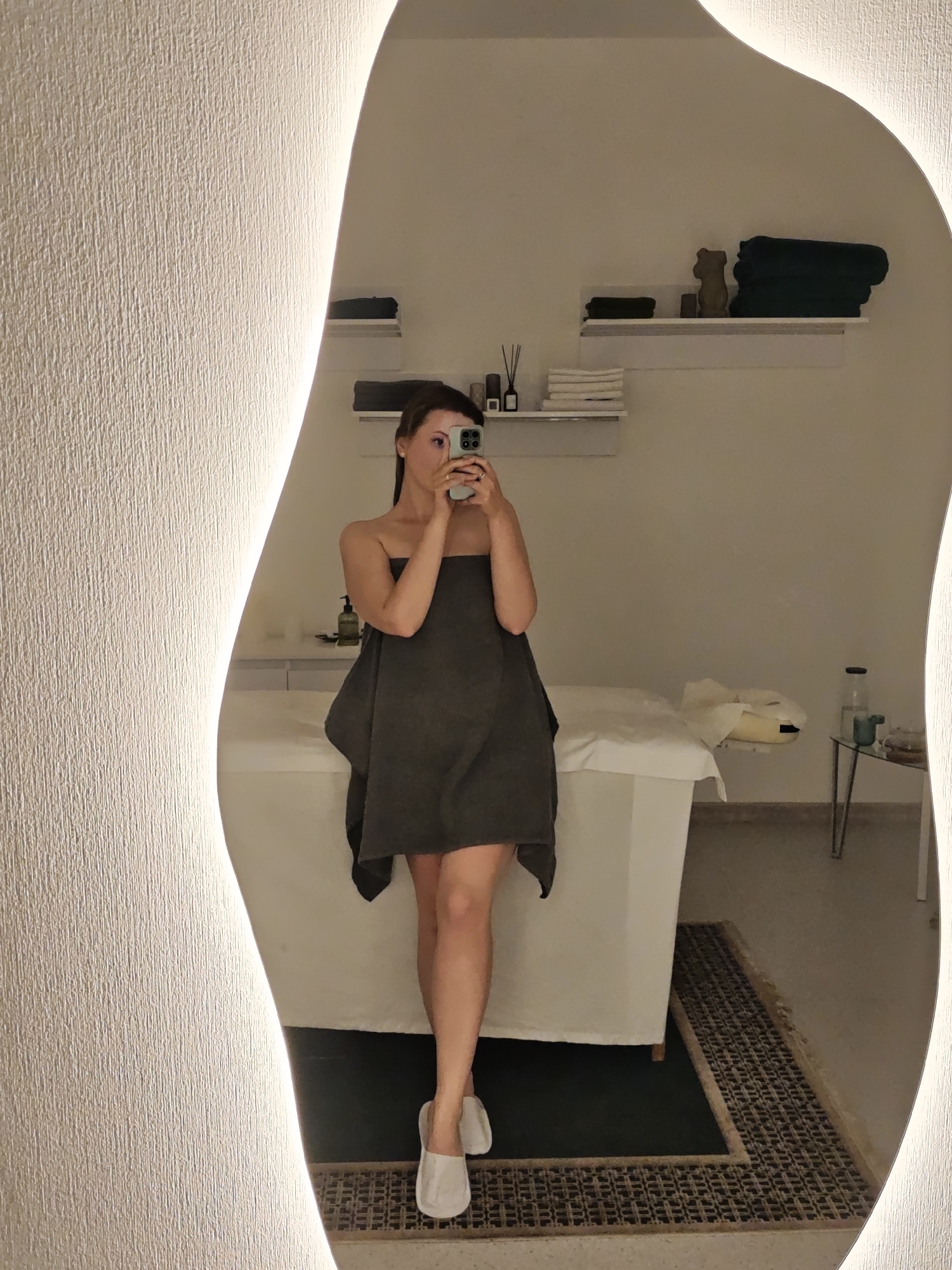 A woman, partially covered in a gray towel and wearing light-colored slippers, takes a selfie in front of a mirror with an irregular, illuminated edge. The mirror reflects a spa or wellness center setting. Behind her is a white massage table and a glass-topped side table holding a bottle and a glass. The background shows shelves with folded towels, a decorative bear, and bottles of liquids or oils. The floor is covered with a dark rug edged with patterned tile. The image was taken in an indoor environment with neutral-colored walls. There is no available information to determine if the picture was taken in Vinnytsia, Ukraine.