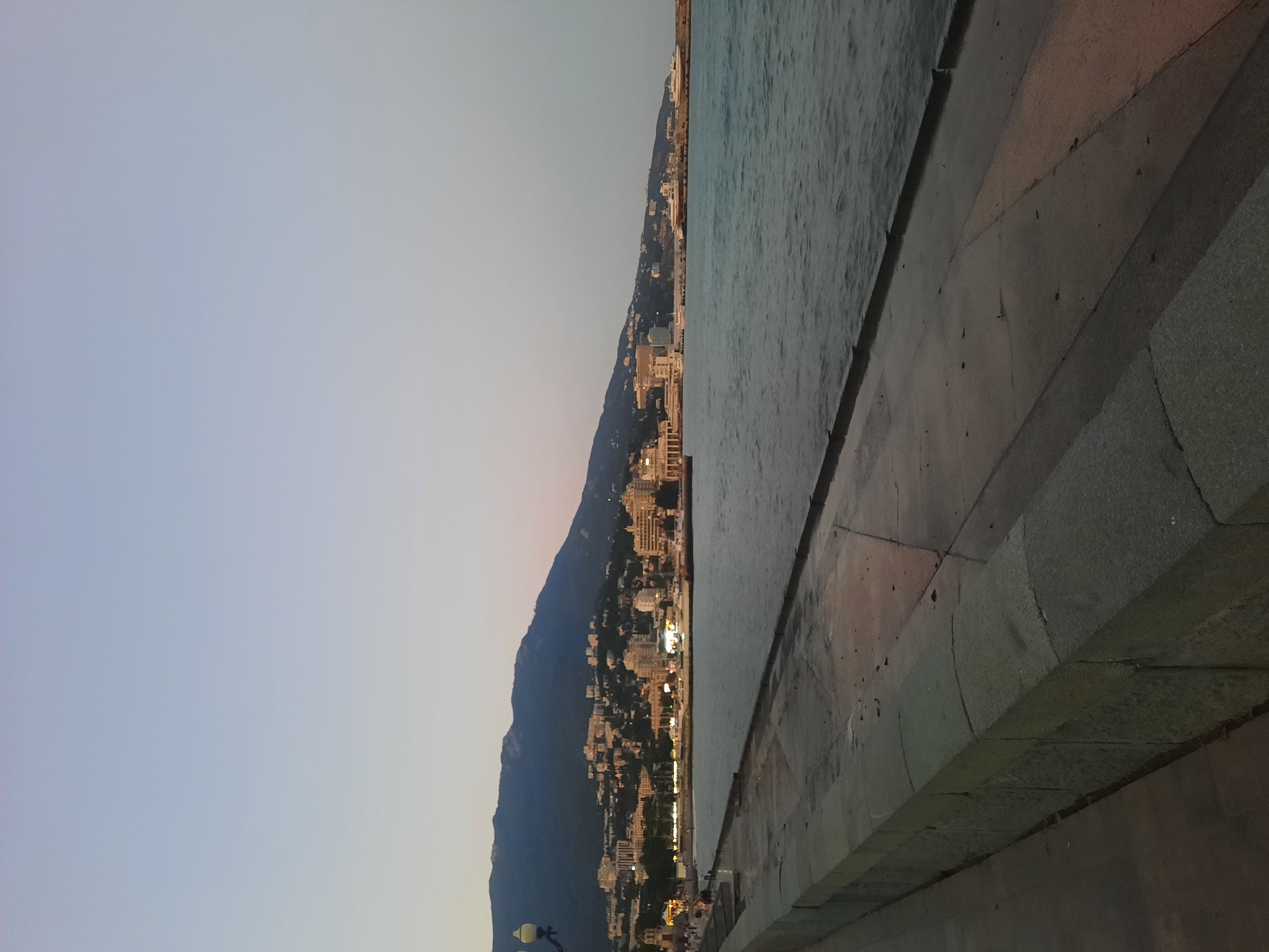 The image depicts a twilight view of the Yalta embankment, a coastal city in Crimea, Ukraine. In the foreground, a stone embankment lines the edge of a body of water, likely the Black Sea, which appears calm with gentle ripples. The embankment is constructed of large, rough-hewn stone blocks. Beyond the embankment, a wide promenade or walkway is visible, paved with concrete and showing signs of wear and possibly moisture.
Further in the mid-ground, the cityscape of Yalta unfolds along the base of a mountain range. Numerous buildings, varying in size and architectural style, are illuminated, indicating the presence of artificial lighting. Some of these buildings appear to be hotels or residential complexes, while others might be public structures. A prominent feature is a breakwater extending into the sea, with several vessels docked or anchored, including what appear to be yachts and possibly a larger ship.
The mountain range in the background is covered in sparse vegetation and is silhouetted against the twilight sky. The sky itself is a soft gradient of pale blue and gray, transitioning towards darker shades at the top, suggesting the time of day is either dawn or dusk. A single lamppost with multiple lights is visible on the left side of the image, near the promenade. A few small figures of people can be discerned walking along the promenade, adding a sense of scale to the scene. The overall atmosphere is one of quietude and natural beauty, characteristic of a seaside city at the end of the day.