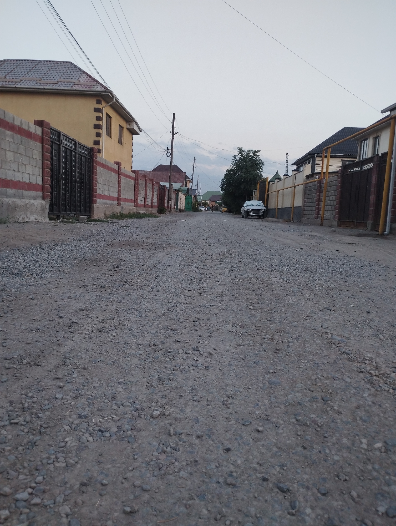 A low-angle, eye-level shot captures a gravel road in a residential area of Novopavlovka, Kyrgyzstan. The road is bordered on both sides by fences and houses. On the left, a yellow house with a dark tiled roof is visible behind a brick and metal fence. The fence features a black metal gate. On the right, a fence with brick posts and a yellow metal railing runs alongside the road, leading to a dark wooden gate. Houses with dark roofs are visible behind these fences. A silver car is parked on the right side of the road in the mid-ground. A single tree stands on the left side of the road in the distance. The sky is overcast and gray, suggesting it is either early morning or late evening. Power lines crisscross the sky above the houses.
