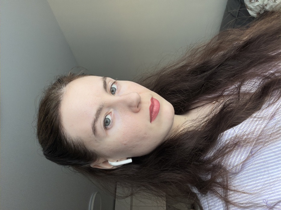 The image displays a head-and-shoulders portrait of an adult female located indoors in what is identified as the city of Ковалёво, Russia. The individual has a light complexion, dark brown wavy hair extending past her shoulders, and blue-grey eyes. She is wearing red lipstick. Visible acne scarring is present on her right cheek. A single white wireless earbud is inserted into her right ear. She is dressed in a light blue and white striped button-up shirt, and a thin gold-colored necklace with a small pendant is visible. Her gaze is directed towards the camera. The background consists of a plain light grey ceiling and a light beige wall. An indistinct dark object is partially visible in the lower right background, and a circular ceiling fixture is partially visible in the upper left background.