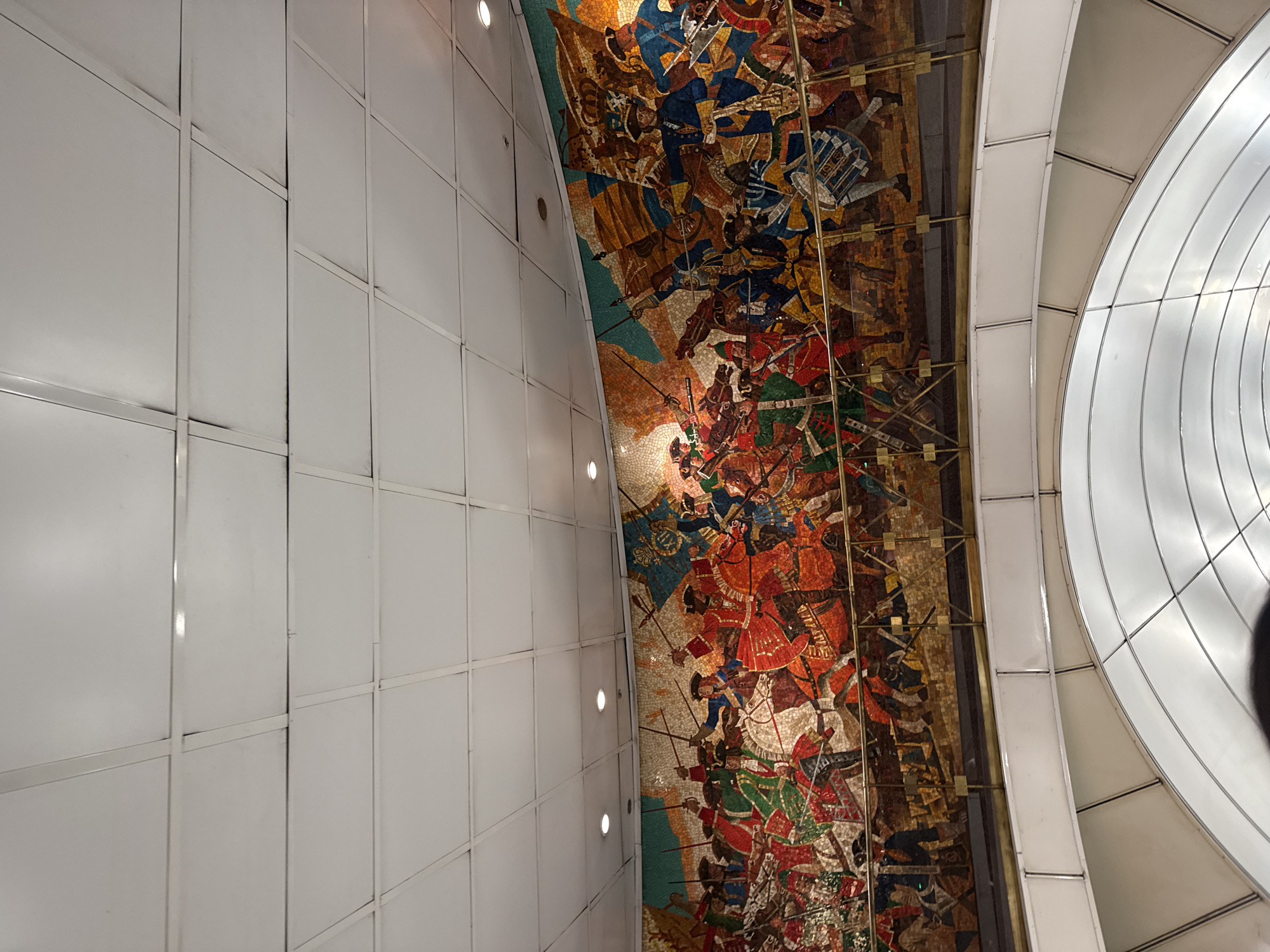 The image displays an upward view within an architectural interior, likely a metro station in Saint Petersburg, Russia. On the left, a tall, white wall is composed of rectangular panels, with several recessed spotlights casting a warm glow.
The central and right portions of the frame feature a large, vibrant mosaic mural adorning a curved ceiling or upper wall. This elaborate mosaic depicts a dynamic historical battle scene. Numerous figures, likely soldiers or warriors, are engaged in combat, some on horseback and others on foot. They are shown in various poses, wielding weapons such as swords and spears. The mosaic uses a rich palette of colors, including prominent reds, blues, greens, oranges, and browns, created from countless small tesserae that give the artwork a textured appearance.
To the far right, a large, white, ribbed, and curved structure forms another part of the ceiling, separated from the mosaic by a slender, possibly metallic or paneled, frame. Artificial lighting from the spotlights illuminates the details of the mosaic and the surrounding architectural elements. The scene conveys a sense of historical narrative and movement. No text is visible in the image.