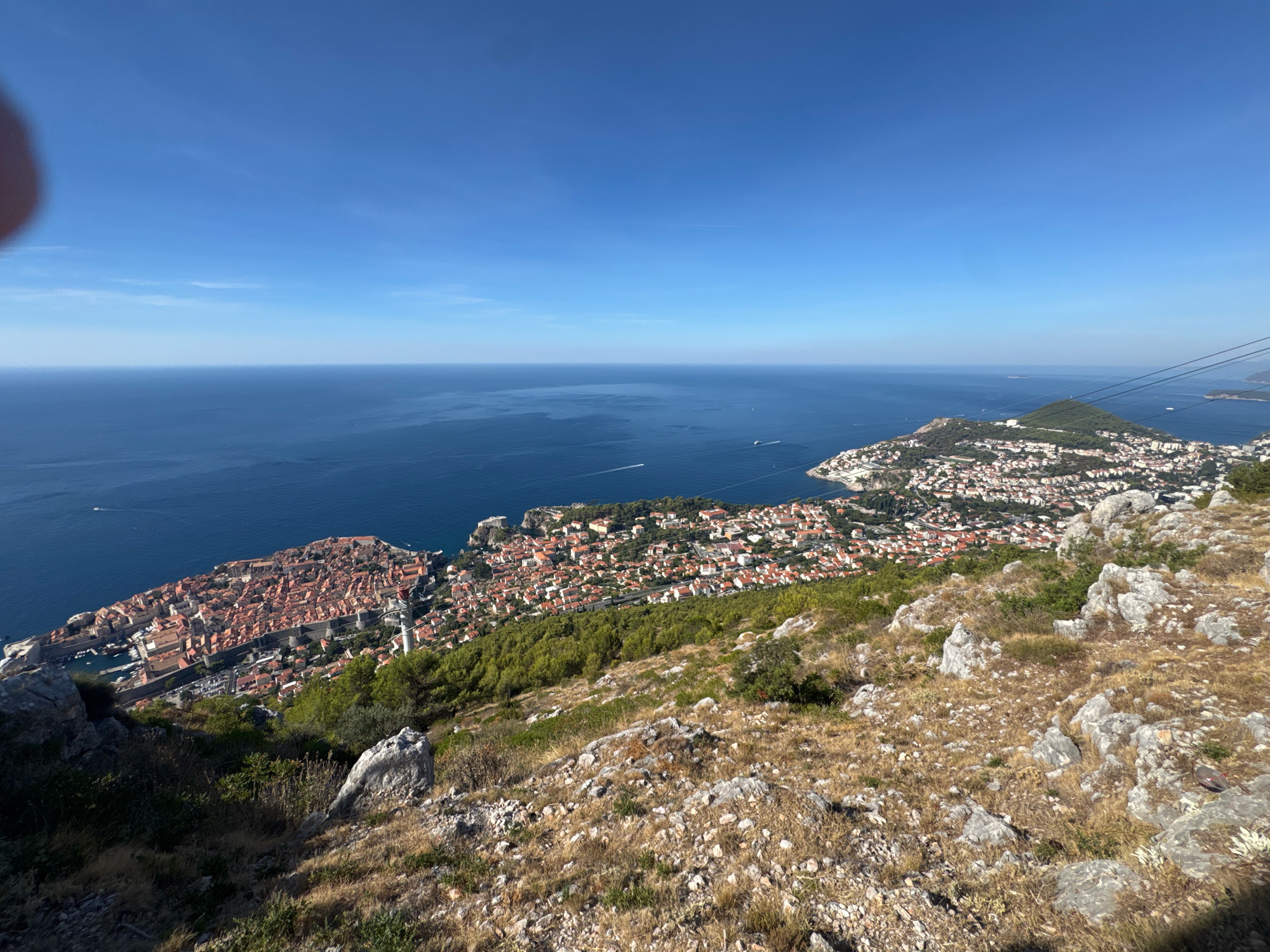 An aerial view captures the historic walled city of Dubrovnik, Croatia, from a high vantage point. The Old Town, characterized by its terracotta rooftops and ancient fortifications, juts into the deep blue Adriatic Sea. The coastline stretches out, dotted with buildings and lush greenery. The foreground is dominated by a rocky, arid hillside with sparse vegetation, suggesting a mountainous or hilly terrain surrounding the city. The sky above is a clear, bright blue, indicating a sunny day. The image appears to have been taken from a cable car or a high viewpoint, possibly Mount Srđ, offering a panoramic perspective of the city and its surrounding landscape.