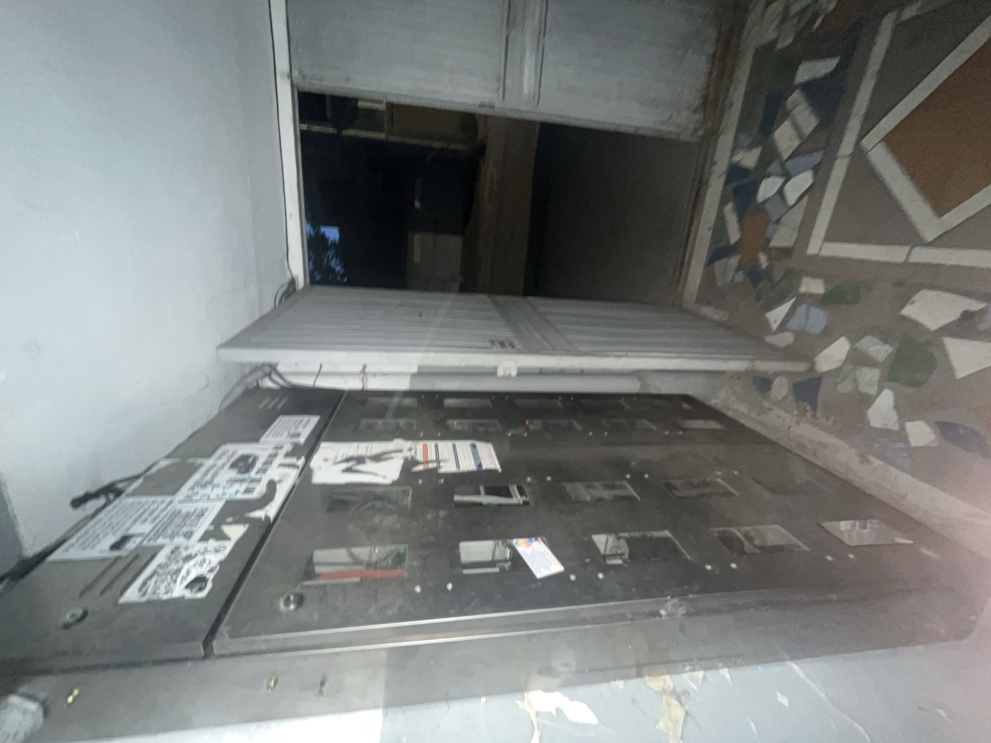 The image shows a close-up of a partially open metal elevator car in Yerevan, Armenia. The elevator door is ajar, revealing a dark elevator shaft. The floor outside the elevator is tiled with a mosaic pattern. The interior of the elevator car has a control panel with buttons and labeling. Some stickers and paper are attached to the control panel. The walls near the elevator show signs of wear and peeling paint. The image appears to be taken from inside the elevator car, looking out towards the building's entryway. No people are visible in the image.