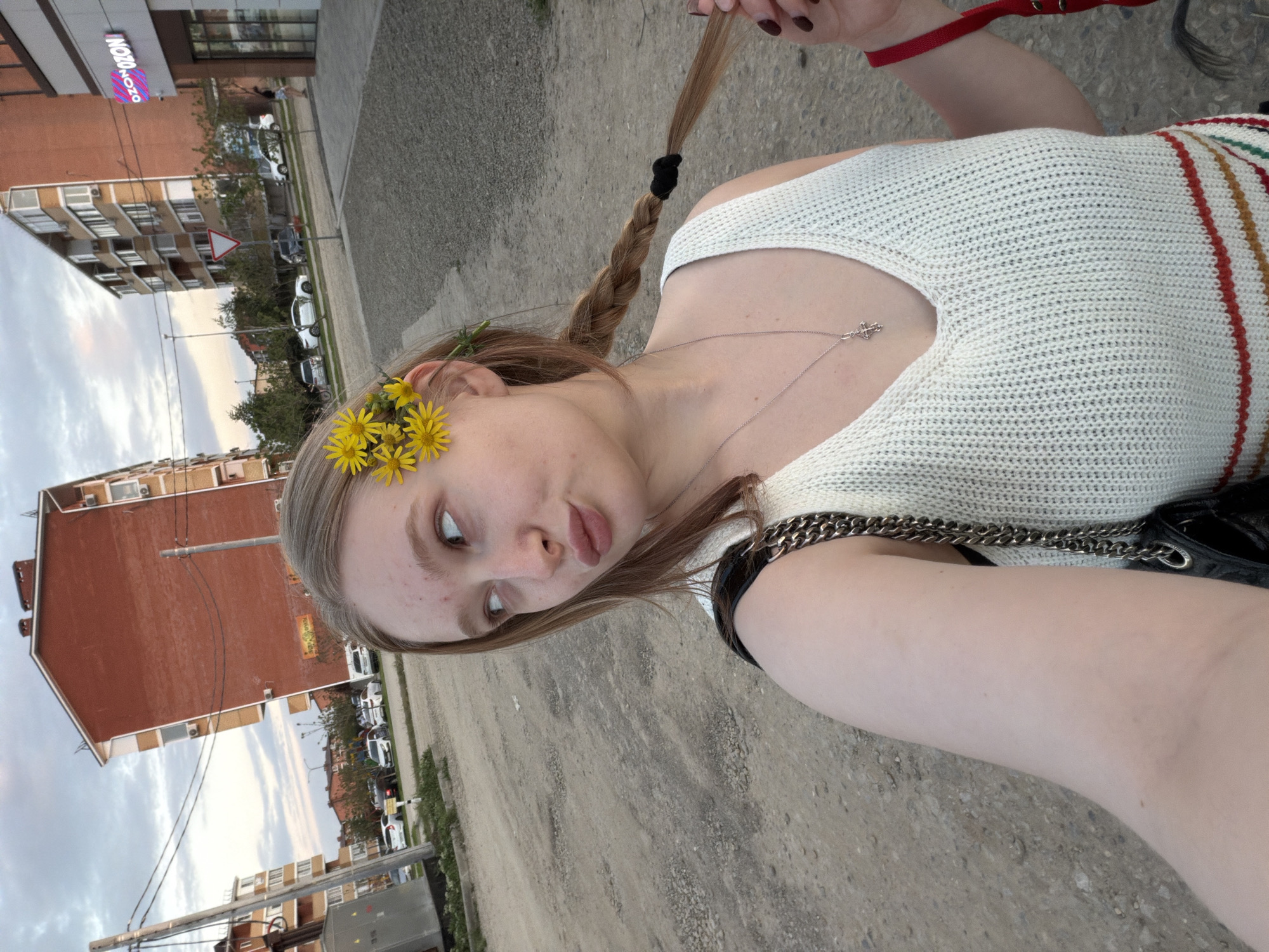 A young Caucasian woman poses for a selfie. Her hair is braided with yellow flowers tucked behind her ear. She wears a white, knit tank top with red and yellow stripes. A silver necklace hangs around her neck. A black purse with a silver chain strap is slung over her shoulder. The woman stands on a dirt and gravel surface. Buildings line the background. A sign written in Cyrillic is visible on a red brick building in the distance. Parked cars are also visible in the distance. Based on the Cyrillic sign and request from the user, the location can be identified as Rossiyskiy, Russia.