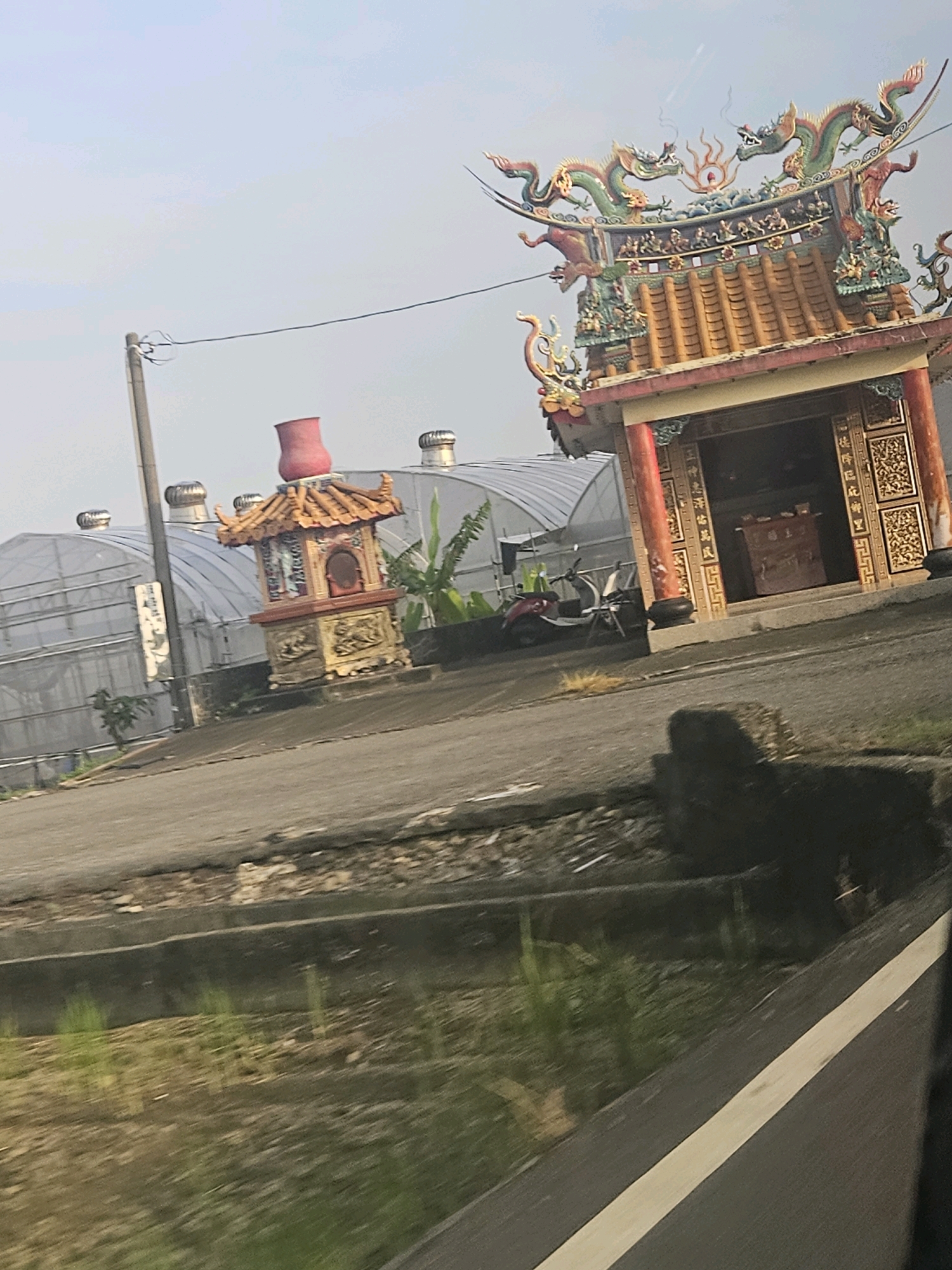 The image captures a view of a traditional Taiwanese temple, possibly located in Xikou Township, Taiwan. In the foreground, a paved road runs diagonally across the frame, with a grassy area and a concrete structure visible on its left side. Further back, a smaller, ornate shrine with a red urn on top stands beside a larger temple. The larger temple features intricate dragon carvings on its roof and decorative pillars. Behind these structures, a row of greenhouses with rounded roofs can be seen, suggesting an agricultural or industrial setting. A scooter is parked to the left of the larger temple. The sky is overcast, with a utility pole and power lines in the background. The overall scene suggests a rural or semi-rural area with a blend of traditional architecture and modern infrastructure.