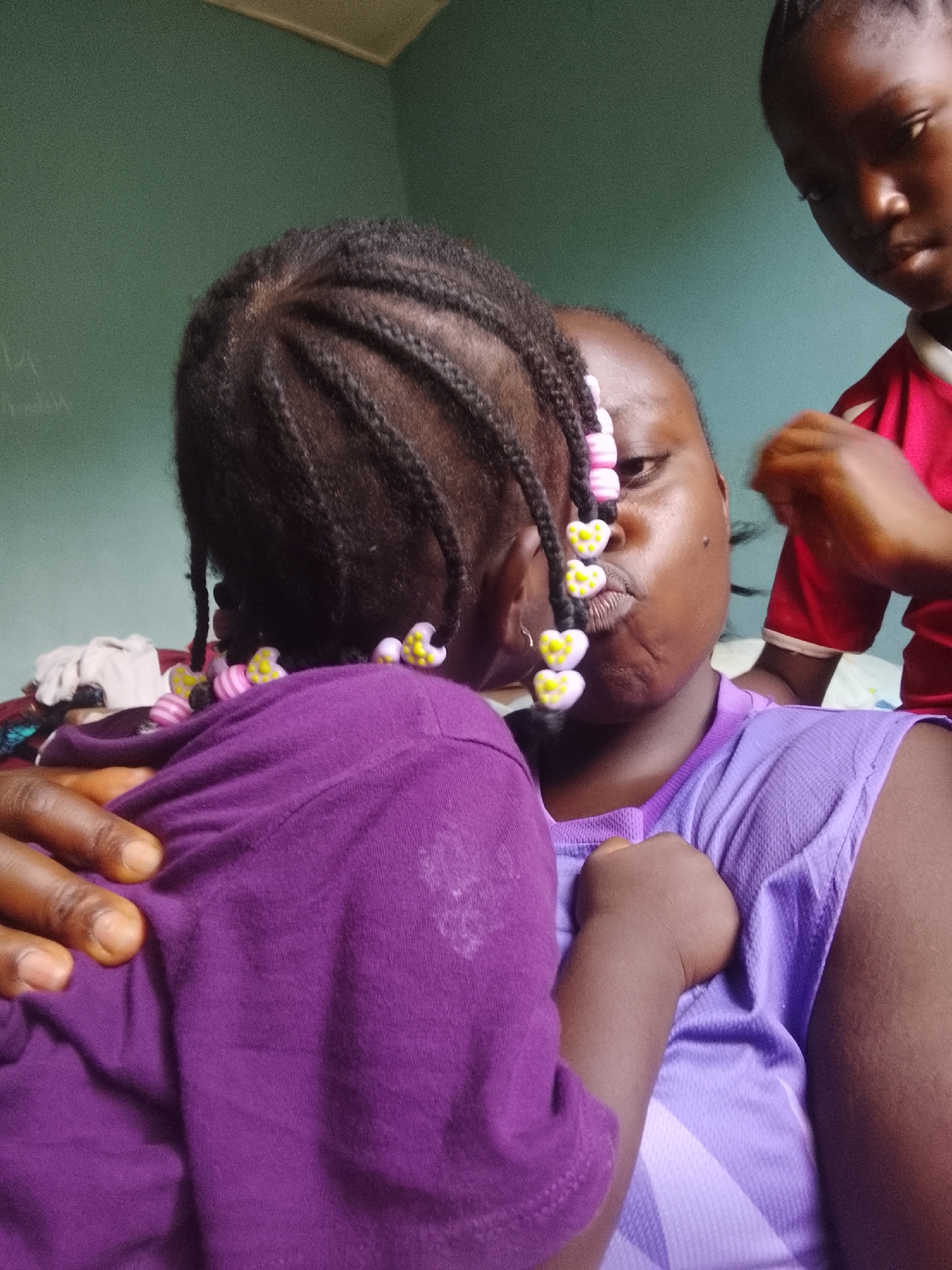 Three individuals are captured in a candid, close-up indoor shot. In the foreground, the back of a young child's head and shoulders are visible. The child has dark hair styled in numerous small braids, many of which are adorned with decorative heart-shaped beads in alternating pink and yellow. They are wearing a long-sleeved purple shirt, and an adult hand is resting on their shoulder, also clothed in purple.
In the midground, an adult female or older youth with dark skin is positioned, facing slightly to the left and looking towards the camera with a playful, "puckered" or "kissing" expression. A few of the same heart-shaped beads are visible near her face, possibly from the child's hair or her own. She is wearing a lighter purple sleeveless top.
Partially visible on the far right is another young person, also with dark skin, whose profile and right arm are shown. Their hand is raised, suggesting they might be styling hair or engaged in another activity. This person is wearing a red and white shirt.
The background consists of a plain, muted green or teal-colored wall. Faint, illegible markings or scribbles are discernible on the upper left section of the wall. The lighting indicates an indoor setting, and the overall mood is intimate and casual. No specific time of day or location cues beyond an indoor room are discernible. There is no visible text.