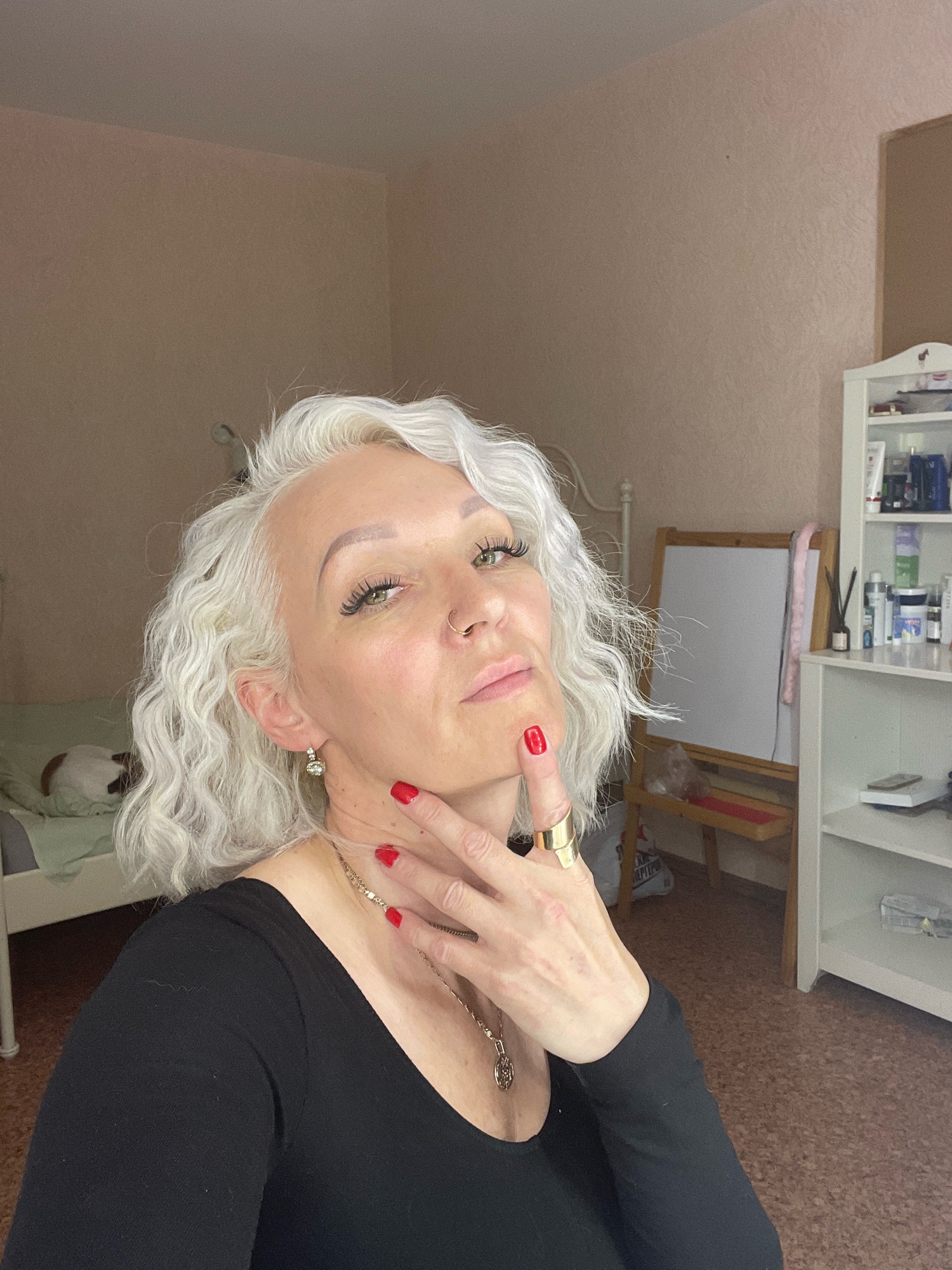 The image shows a woman with white, wavy hair and green eyes, wearing a black top and red nail polish. She is indoors, in what appears to be a bedroom or living space. A bed with a light green duvet is visible in the background to the left, with a white headboard. To the right, there is a white bookshelf filled with various items, and an easel with a blank canvas. The lighting suggests it is daytime, with soft light coming from an unseen window. The overall impression is a candid, personal moment captured at home. There is no indication of the location being in Saint Petersburg, Russia.