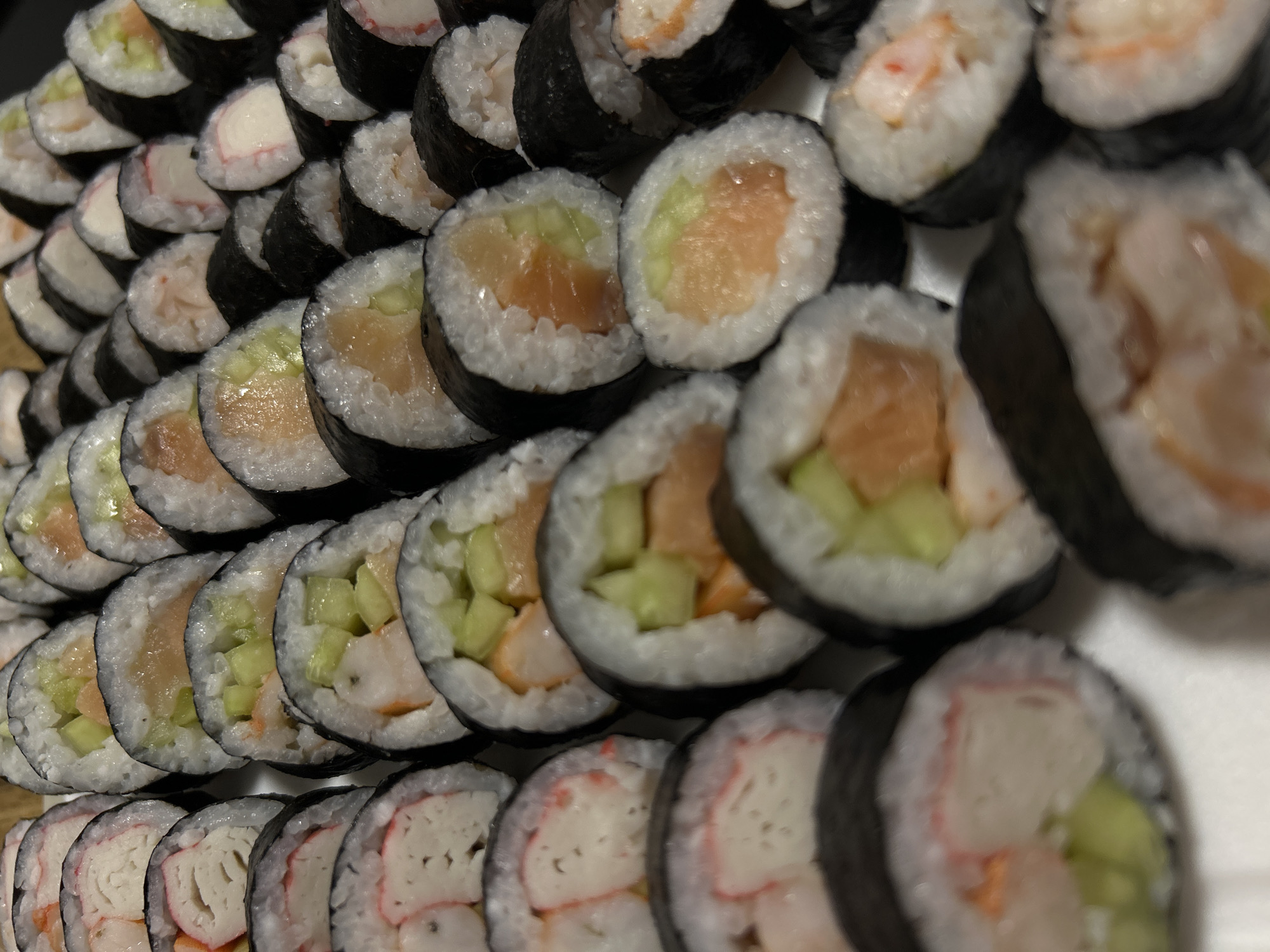 A high-angle, close-up shot shows a large assortment of sushi rolls arranged on a white surface. The rolls are of varying ingredients, including what appears to be imitation crab, salmon, and cucumber. The sushi is cut into bite-sized pieces and arranged in a dense, grid-like pattern. The lighting is even, and there are no visible shadows. The setting is indoors, with no discernible urban or natural elements. This image does not contain any information to suggest a location in Szczecin, Poland.