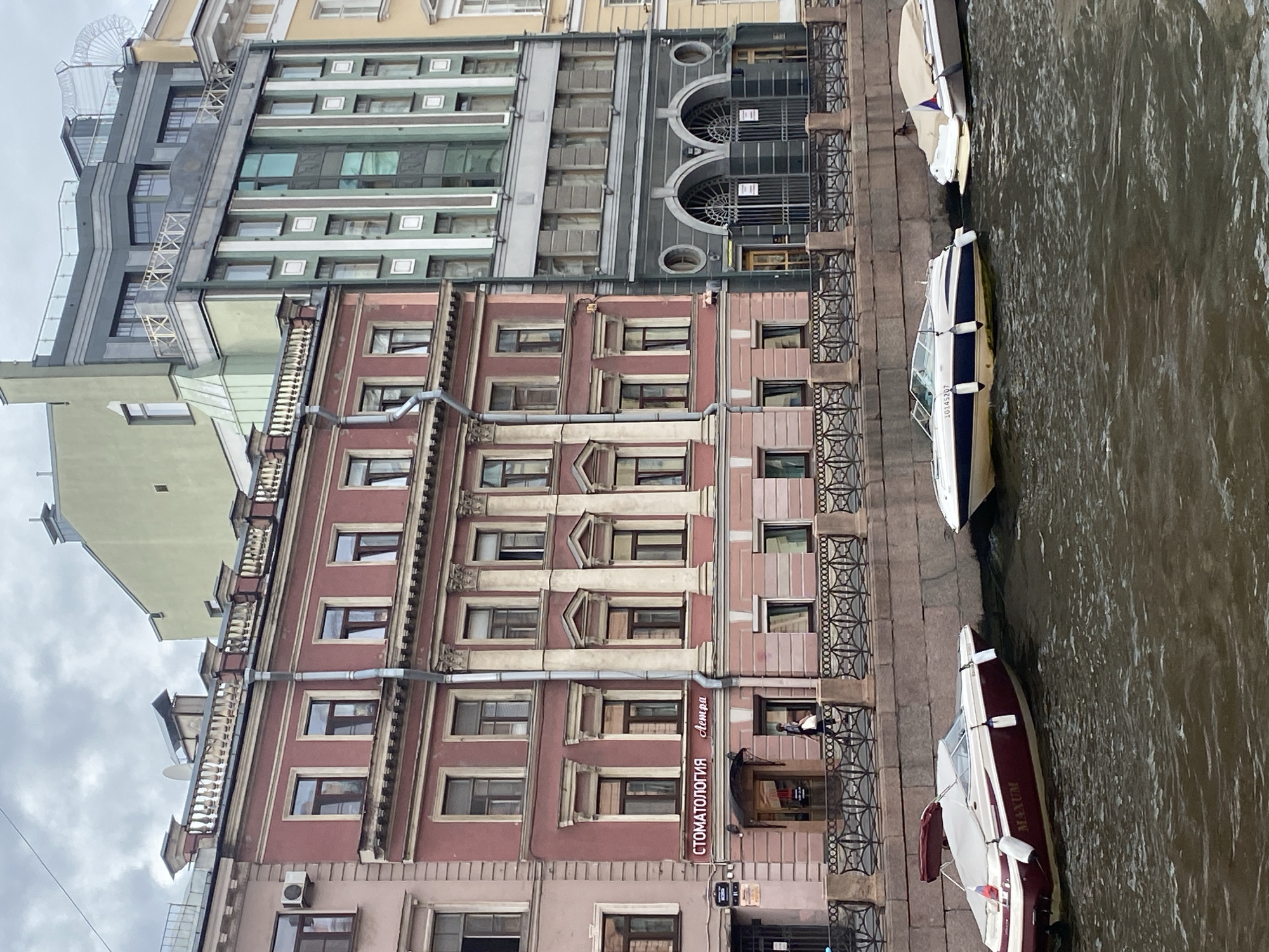 This is a photograph taken in Saint Petersburg, Russia, likely in the historic center, depicting a canal-side scene. The foreground shows several pleasure boats moored in the dark, rippling water of a canal. The boats are of varying sizes and styles, with one prominent burgundy and white motorboat bearing the name "MAXUM" and a Russian flag on its stern. To the right, another white boat is partially covered.
The middle ground features a quay lined with a decorative metal fence. Behind the fence are several multi-story buildings. The most prominent building on the left is a pink, ornate structure with classical architectural details, including columns and arched windows. A sign on this building reads "СТОМАТОЛОГИЯ Астра" (Dentistry Astra). A person dressed in dark clothing is walking along the quay in front of this building.
Adjacent to the pink building is a modern, grey and green building with a more contemporary design, featuring large windows and geometric patterns. Further to the right, a lighter-colored building with yellow accents is visible. The overall atmosphere suggests an overcast day, with a muted color palette. The buildings appear to be old and well-maintained, reflecting the architectural heritage of Saint Petersburg. The canal itself is a common feature of the city, often referred to as the "Venice of the North."