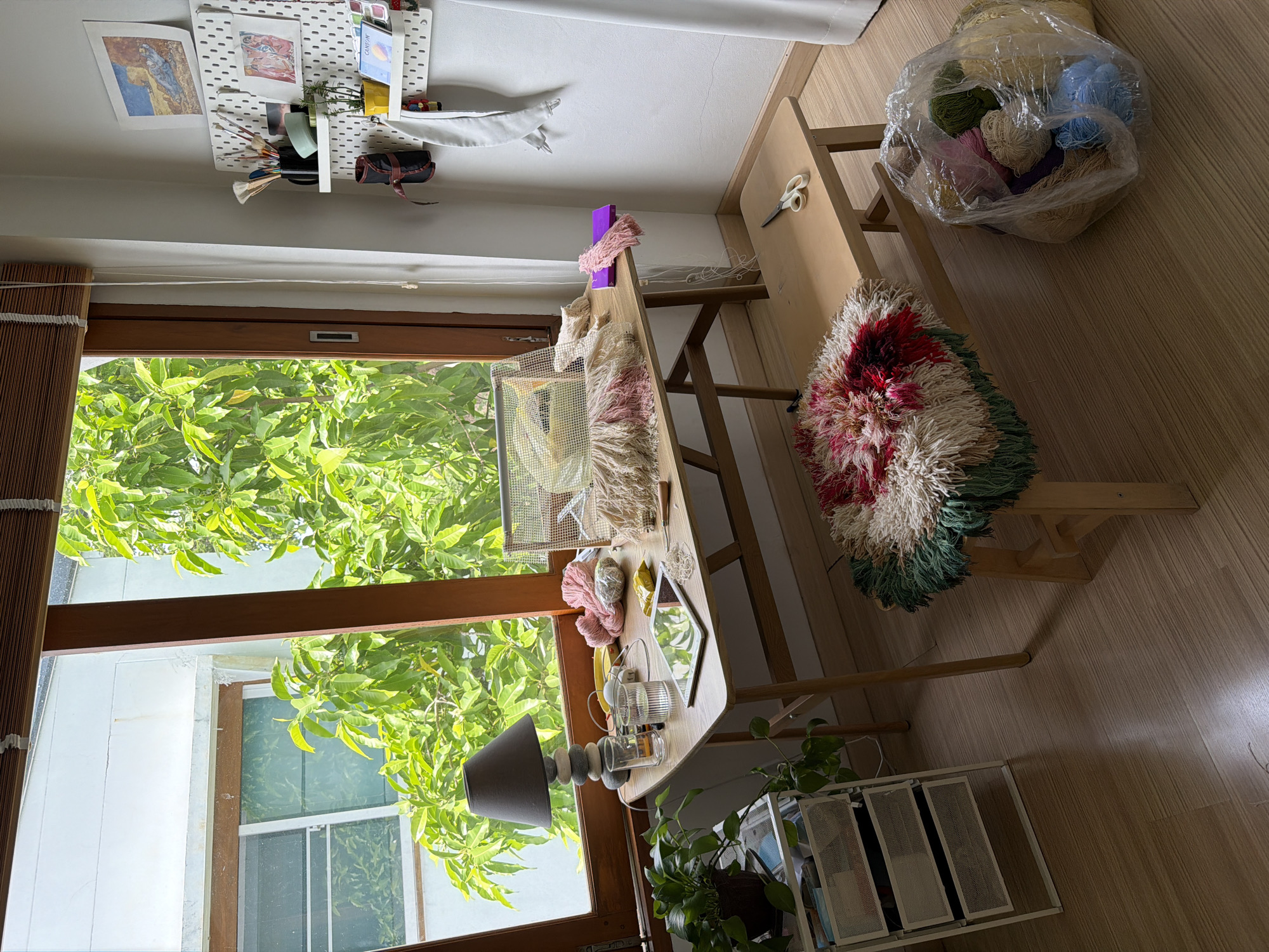 This is a vertical, medium-shot, interior photograph taken in what appears to be a craft or art studio in Chang Phueak, Thailand. The room is well-lit, suggesting daytime. Sunlight streams in from a large window, illuminating the wooden floor and casting shadows.
The main subject is a wooden desk with various craft supplies. On the desk, there are balls of yarn in various colors, a pair of scissors, a small mirror, some glassware, and a table lamp with a grey shade. A partially finished weaving or rug with colorful fringes is draped over a wooden stool next to the desk. A large plastic bag filled with more balls of yarn is visible on the floor next to the stool.
The window provides a view of lush green foliage outside, indicating a tropical or sub-tropical climate. Beyond the foliage, parts of a white building and another window are visible.
On the wall above the window, there is a white pegboard system adorned with artwork, some small plants, and art supplies like brushes. The overall impression is of a creative and organized workspace. The lighting and the presence of plants suggest a relaxed and natural ambiance. There is no visible text in the image.