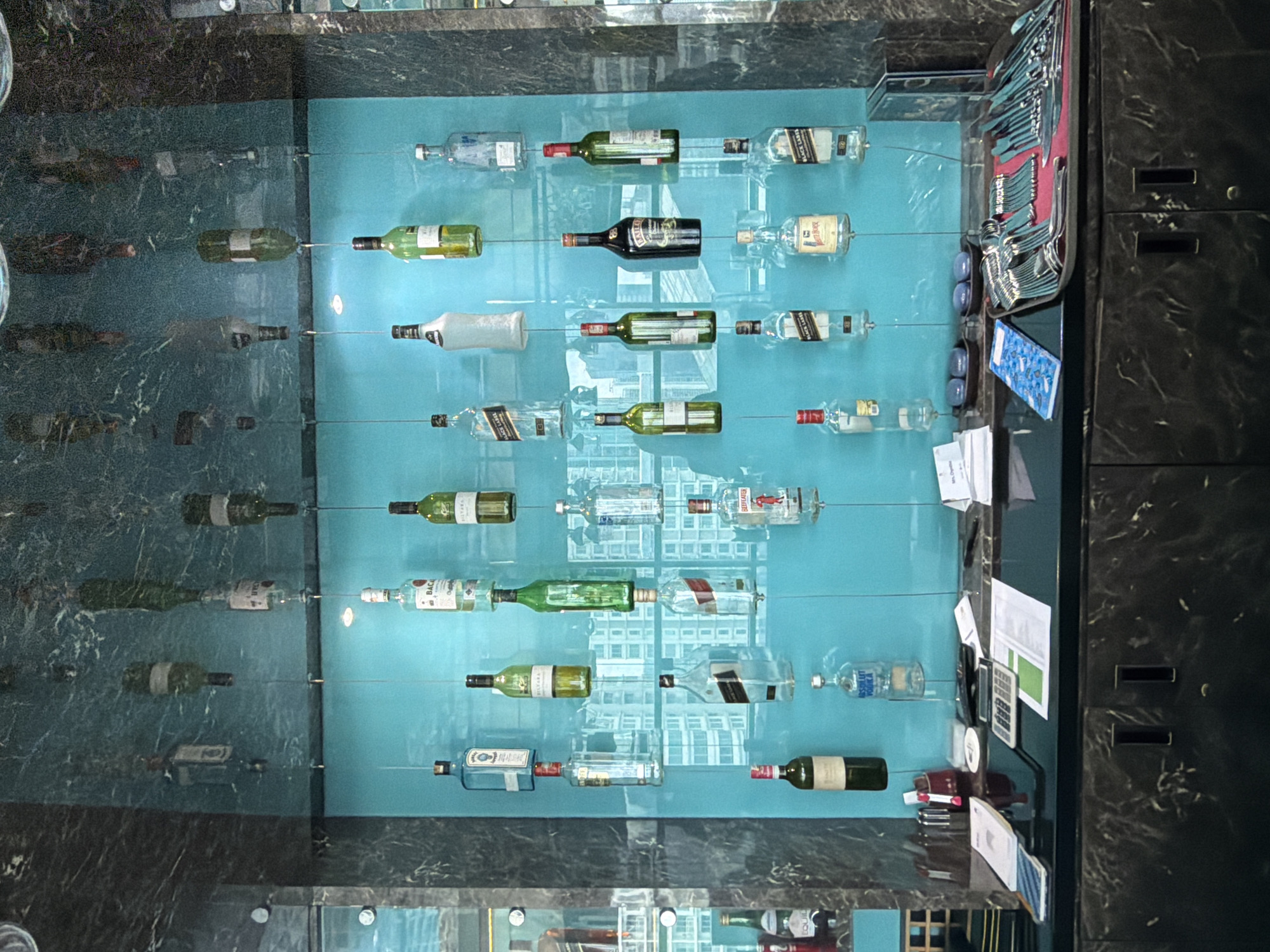A display of liquor bottles is arranged horizontally on a turquoise surface, likely in a bar or restaurant setting. The bottles are mostly empty and appear to be supported by thin wires or rods, giving the impression of them floating in the air. Some bottles are green, while others are clear.
To the right of the liquor display is a set of what appears to be cutlery, including forks and spoons. Alongside the cutlery are some office supplies such as pens, calculators and loose pieces of paper. The wall is decorated in a dark, marbled pattern and looks like a cabinet.
The location is South Jakarta, Indonesia.