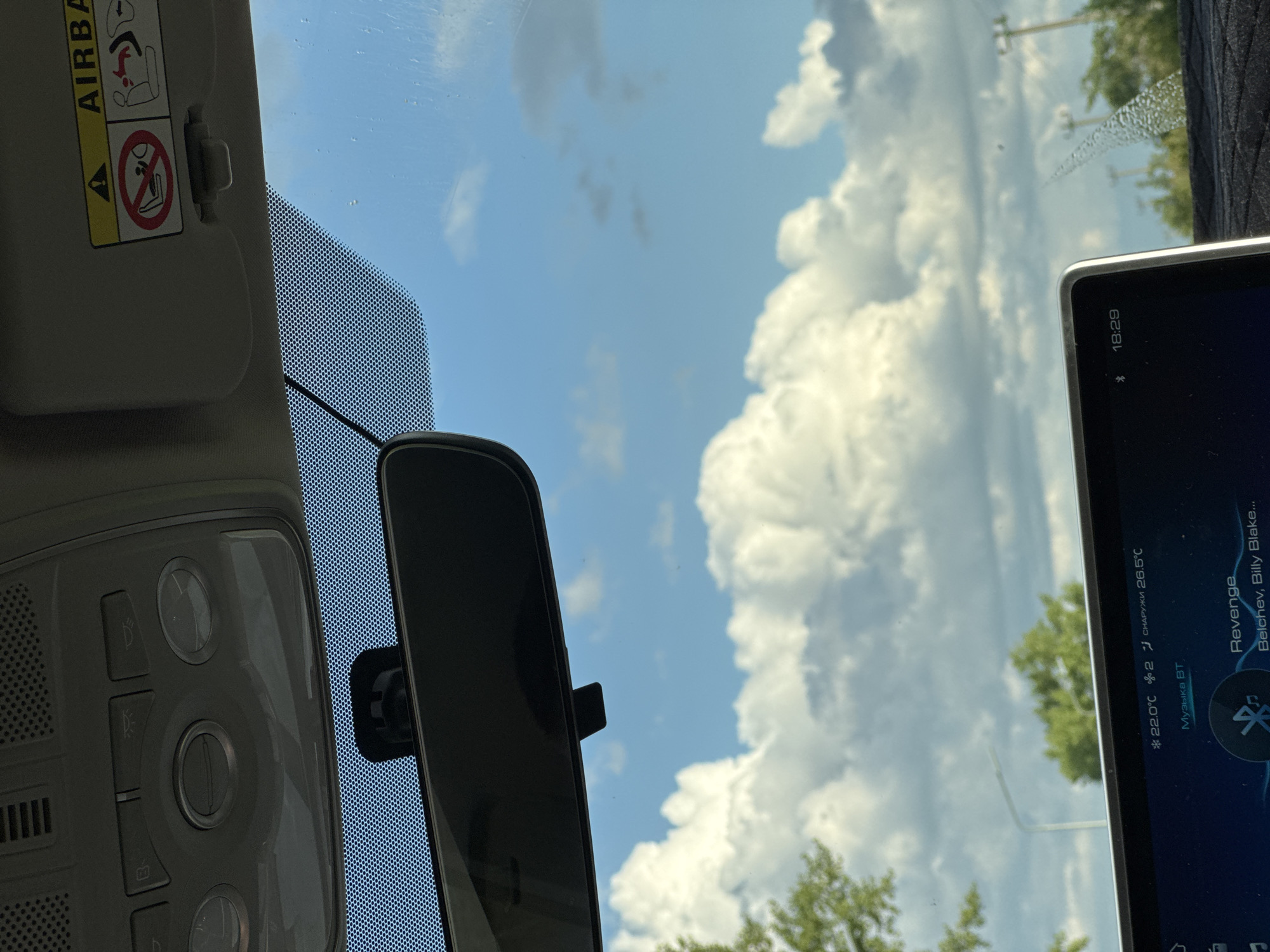 The image shows the interior of a car looking out through the windshield at a bright blue sky filled with fluffy white clouds. The car's rearview mirror is visible, as is the car's overhead console with various buttons and lights. A sun visor with safety warnings, including an airbag symbol and a graphic indicating no children in the front seat, is also present. The car's dashboard display screen shows the time as 18:29 and song information, including the title "Revenge" by Belchev, Billy Blake. Temperature readings of 22.0°C inside and 26.5°C outside are also displayed. The location is identified as Novomar'evskaya, Russia. Trees and a street light are visible in the background outside the car.