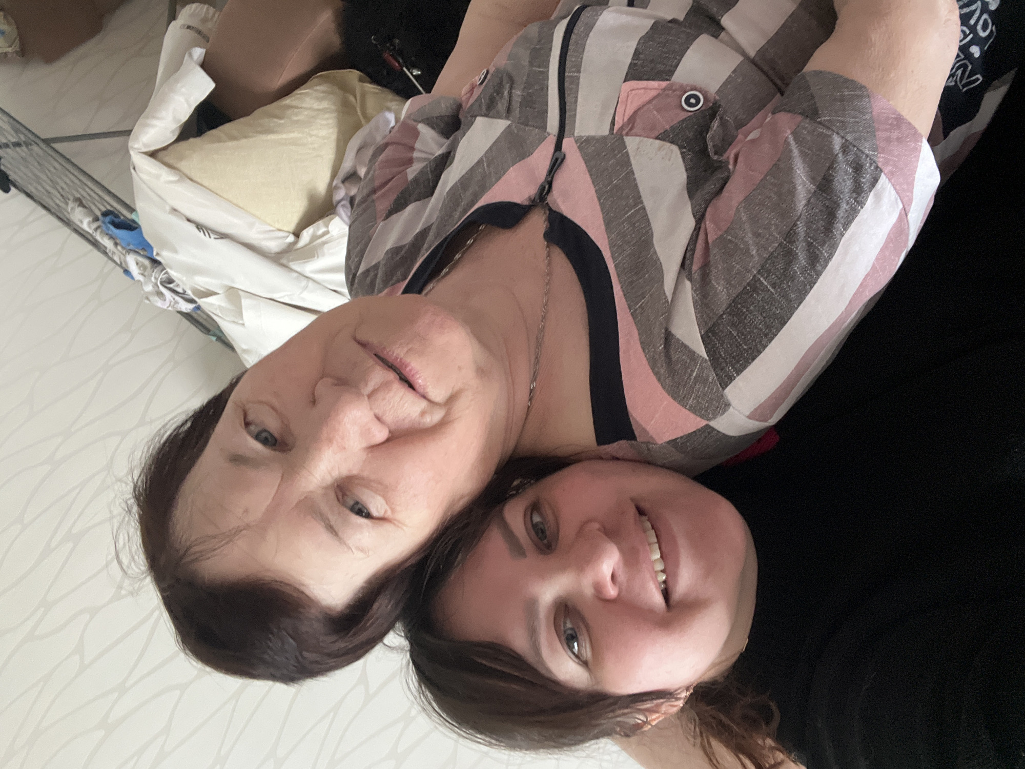 The image captures two women, likely family members, posing closely together in an indoor setting in Perm, Russia. The photograph is rotated 90 degrees clockwise.
The woman on the upper side of the frame (appearing older) has dark brown hair, blue eyes, and a neutral expression with a slight smile. She is wearing a button-up shirt with a distinctive striped pattern in pink, grey, and white. A silver chain necklace is visible around her neck.
The woman on the lower side of the frame (appearing younger) has dark brown hair, blue eyes, and is smiling broadly, showing her teeth. She appears cheerful and is wearing a dark, possibly black, top. On her black clothing, faint white text is partially visible, reading "LOVE" and "WN" with the rest cut off.
They appear to be lying down or leaning back, with their heads close together, looking towards the camera. The background features a light-colored wall with a subtle, wavy textured pattern. Behind the older woman, elements of a bed or couch are visible, including pillows and white bedding. One pillow is a pale yellow. A metal grid-like structure, possibly a shelf or part of a bed frame, is also seen on the left. The lighting suggests it is daytime. The overall impression is one of warmth and affection.