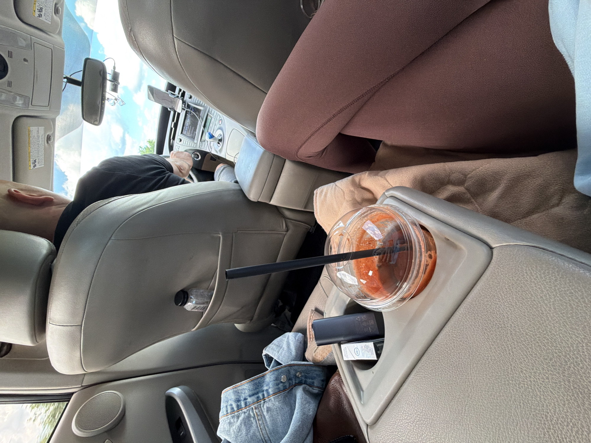 The image depicts the interior of a car, likely a hybrid vehicle given the center console. In the foreground, a clear plastic cup filled with an orange beverage and a black straw sits in a cupholder. Next to it are two small black electronic devices, possibly a vape pen and a charger, and a white rectangular package, likely for cigarettes or similar products. A denim jacket is draped over the armrest. The driver, a bald man wearing a black t-shirt, is visible in the driver's seat, his hands on the steering wheel. The car appears to be in motion, with a view of the sky and trees through the windows. The interior is primarily composed of beige and gray leather seats and plastic components. The presence of a small water bottle in a seat-back pocket on the passenger side suggests a journey. The image is taken from the perspective of someone sitting in the back seat. No specific landmarks or indicators of Step-Soci, Moldova are present in the image.