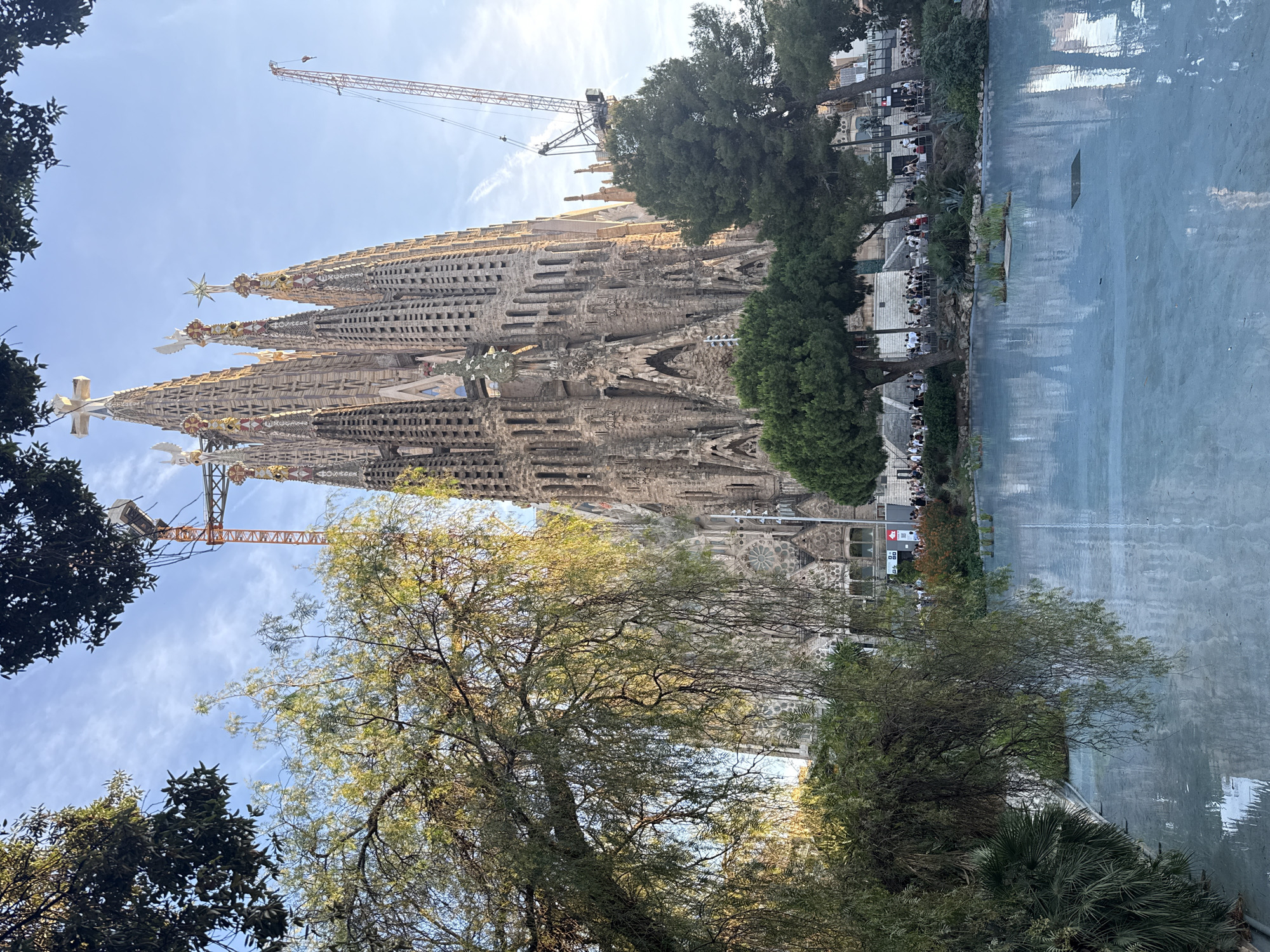The image depicts the Sagrada Familia, a Roman Catholic minor basilica in Barcelona, Spain, viewed from across a pale blue, shallow pool of water. The basilica's iconic, unfinished towers dominate the background, with a large orange construction crane positioned to the right of the structure. Lush green trees, including a prominent weeping willow-like tree in the foreground left, frame the scene. The sky is a clear blue with wispy white clouds. In the middle ground, behind the pool, a group of people are visible walking along a pathway in front of the basilica, with more trees and landscaping. The pool's surface reflects the sky and the surrounding architecture.