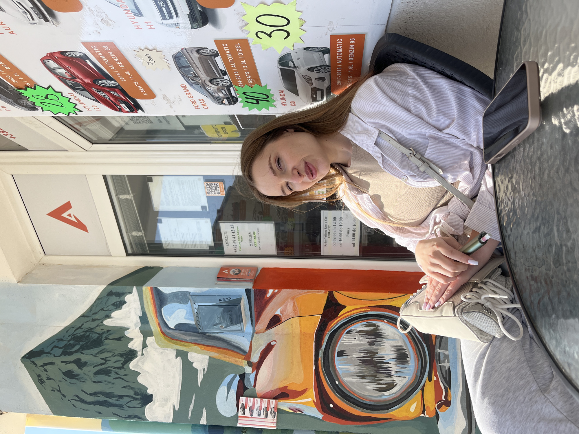 A female individual is seated outdoors on a dark chair at a glass-top table, positioned in front of a car rental agency. The location is identified as Budva, Montenegro, based on the displayed Montenegrin country code (+382), the company name "Rent a Car ADRIATIC," and associated websites (GETCAR.ME, RENTACARADRIATIC.COM), which commonly operate in this tourist city.
The woman has light brown hair and is dressed in a light-striped button-up shirt over a beige top and light grey sweatpants. She is looking towards the camera with her left eye partially closed and lips slightly pursed. Her left leg is bent, and she holds a light-colored sneaker with both hands. A small, dark-colored electronic device, possibly a vape, is visible in her right hand near the shoe. A gold-colored smartphone with a dark screen rests face-up on the table in front of her.
To her left, a colorful mural on a building pillar depicts a green mountain, white clouds, a rainbow, and the front section of an orange and yellow car featuring a prominent circular headlight. A metal key drop-box is affixed to the pillar.
To her right, the building's white facade features glass doors and windows displaying multiple car rental advertisements. These ads show various car models, including a red Hyundai i20 (2014, automatic, 5 seats), a brown Ford Grand C-Max (2014-2018, automatic, 7 seats), and a white Hyundai i30 (2017-2018, automatic, 5 seats), along with prices (e.g., 28, 40, 30) and specifications. The agency's red 'A' logo is prominent. Contact information, including a Montenegrin phone number (+382 69 41 42 43) and specific working hours, is displayed. The scene is illuminated by bright daylight.